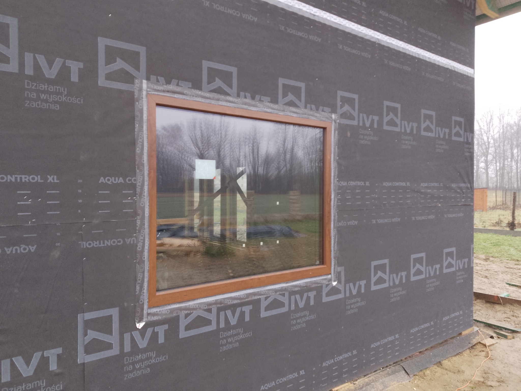 Montaż okna PCV z brązową ramą w budynku z czarną membraną izolacyjną. Widoczne uszczelnienia i taśmy izolacyjne. W tle drzewa bez liści.