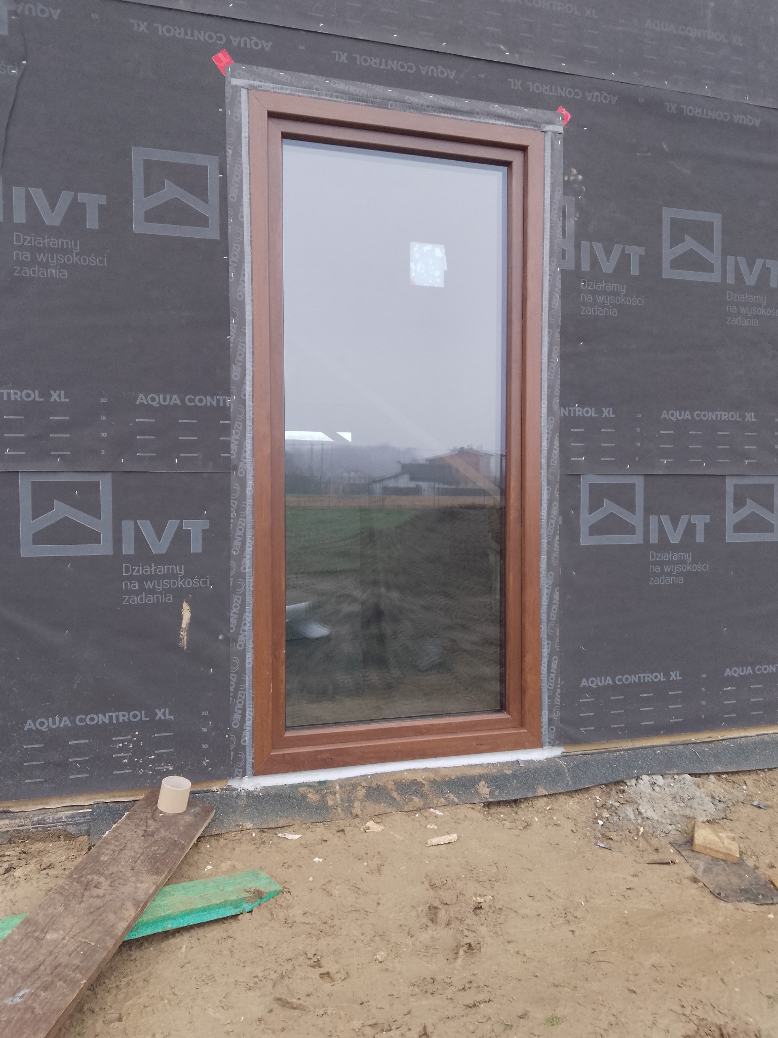Brązowe okno PCV zamontowane w surowym stanie budynku, widoczne folie izolacyjne i fragmenty desek na ziemi. Ujęcie z bliska, podkreślające detale montażu.