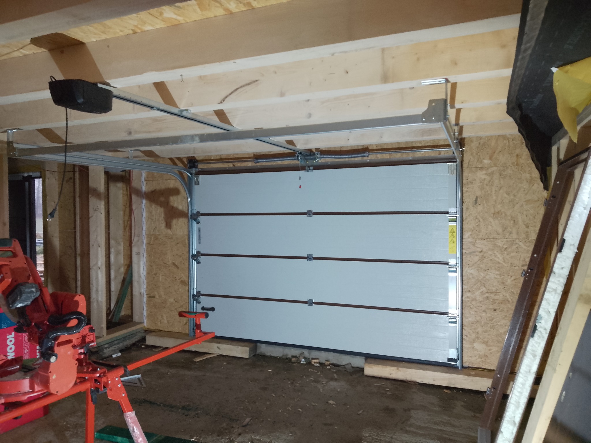 Montaż segmentowej bramy garażowej z napędem, widoczne prowadnice i konstrukcja nośna. W tle surowa konstrukcja garażu z drewnianych belek.