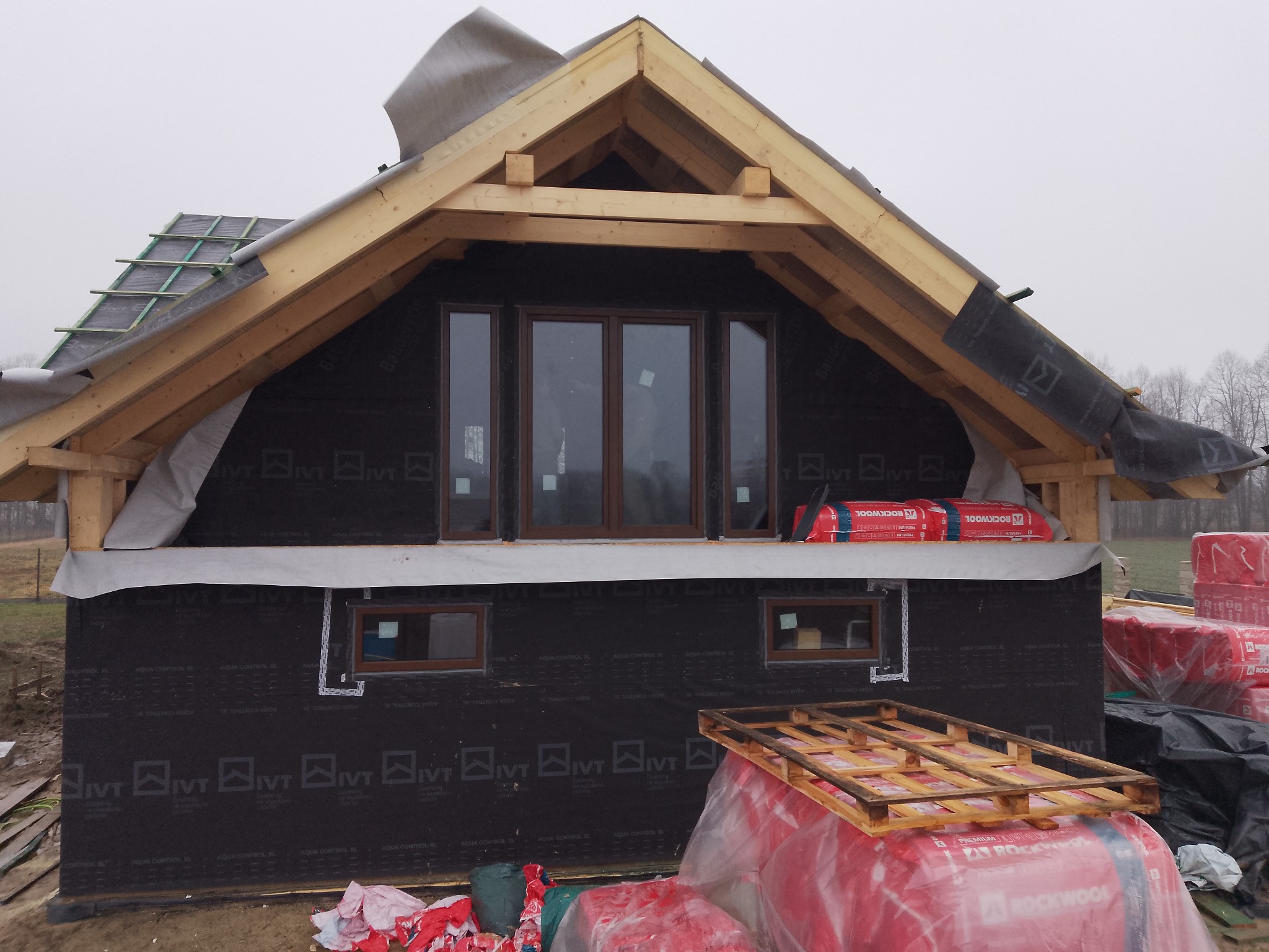 Budowa domu w trakcie: widoczne okna z brązowymi ramami, drewniana konstrukcja dachu, czarna membrana i ocieplenie Rockwool. Etap prac wykończeniowych.