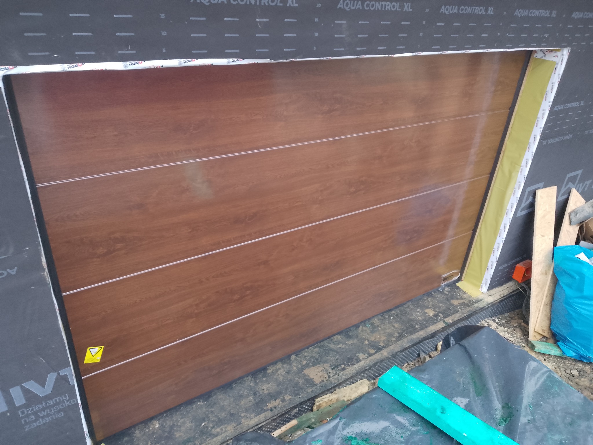 Montaż segmentowej bramy garażowej w kolorze drewna, widok z bliska na proces instalacji, z widocznymi elementami konstrukcyjnymi i izolacją.