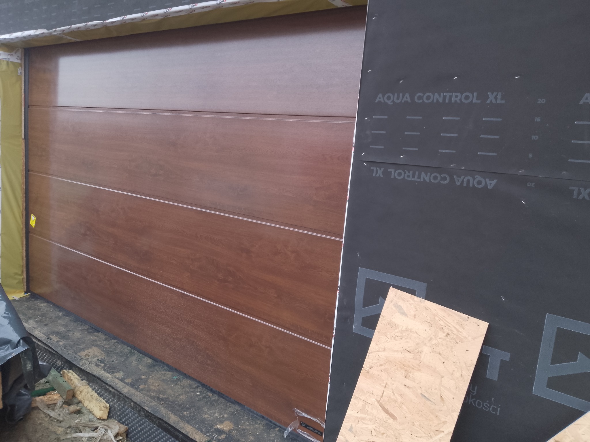 Montaż segmentowej bramy garażowej w kolorze drewna, widok z boku, z widoczną folią ochronną i elementami montażowymi. Detal wykończenia.