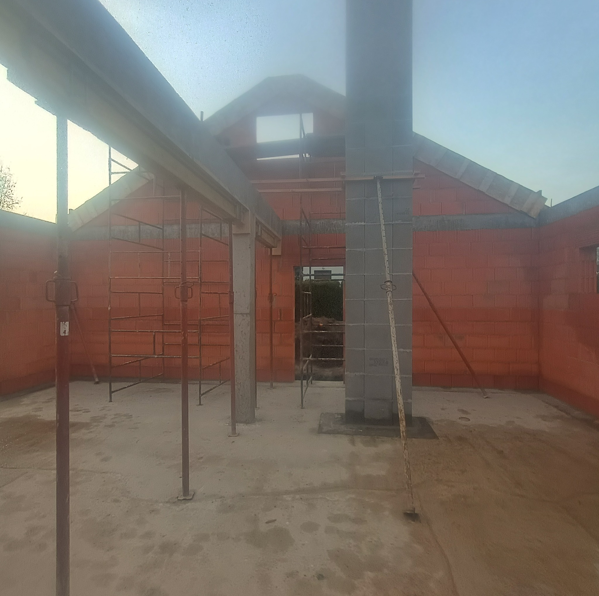 Wnętrze budynku w trakcie budowy z czerwonej cegły, widoczne rusztowania, betonowe słupy i komin z szarych bloczków. Surowy stan, przygotowanie do dalszych prac.