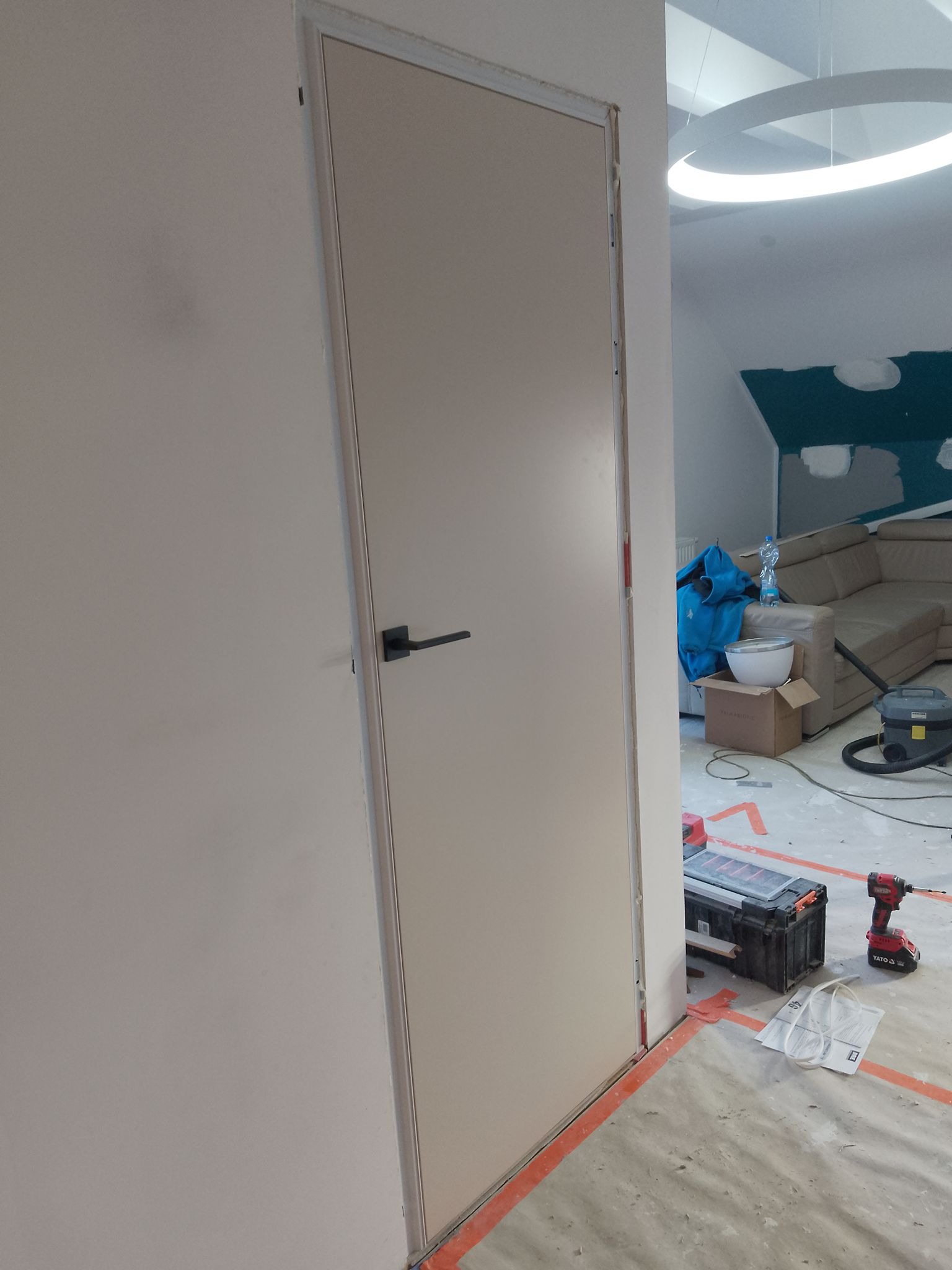 Zamknięte, beżowe drzwi wewnętrzne z czarną klamką, widoczne narzędzia i elementy wykończeniowe w tle. Montaż drzwi w trakcie remontu.