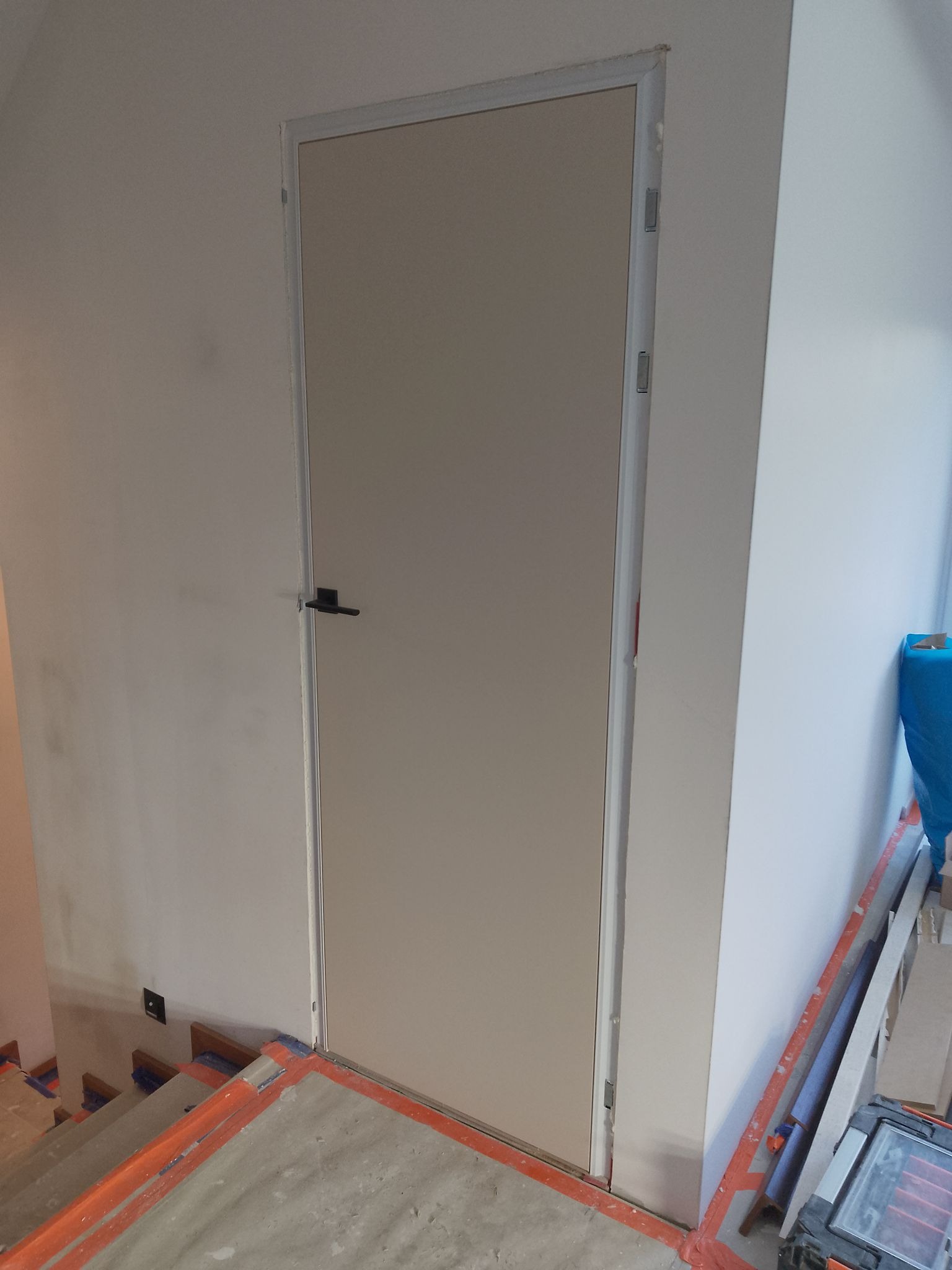 Zamknięte, beżowe drzwi wewnętrzne z czarną klamką, osadzone w białej futrynie. Widoczne zabezpieczenia na schodach i podłodze, sugerujące prace wykończeniowe.