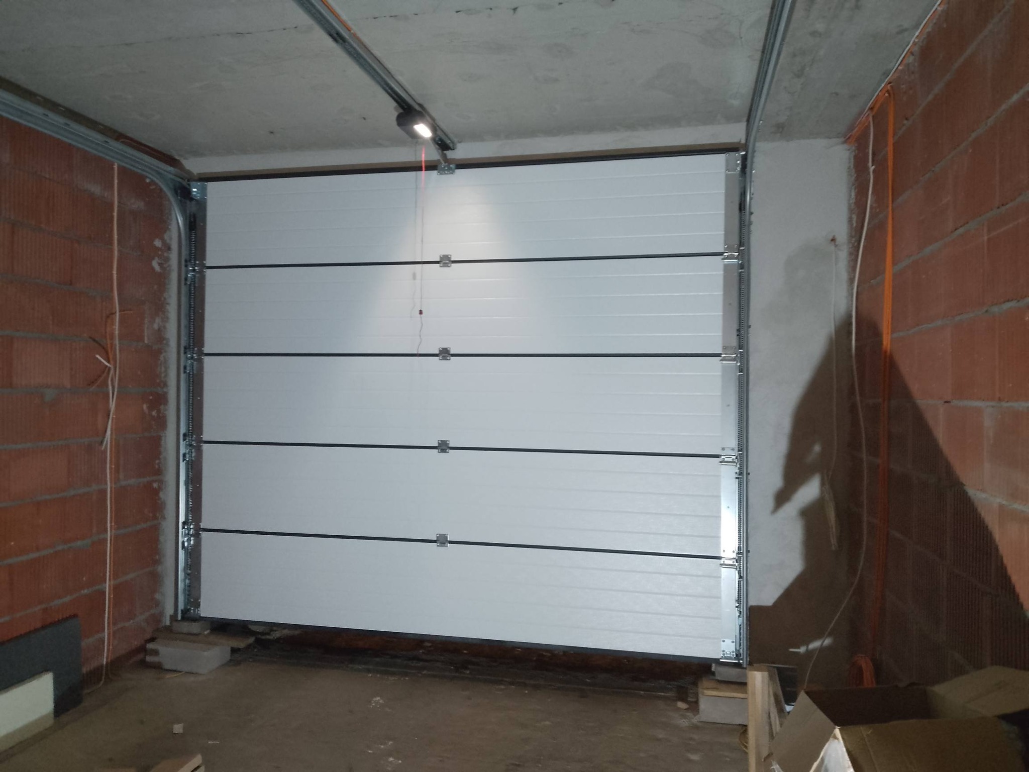 Zamknięta, segmentowa brama garażowa w surowym garażu z ceglanymi ścianami i betonowym sufitem. Widoczne prowadnice i mechanizm bramy.