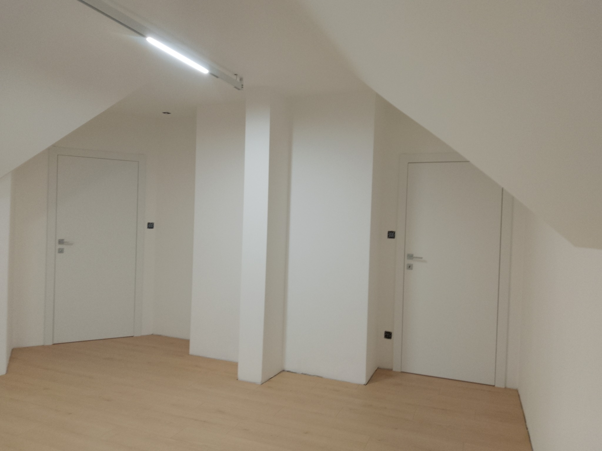 Dwie białe, minimalistyczne drzwi wewnętrzne w skosach poddasza, z widoczną podłogą w odcieniach drewna i białymi ścianami. Oświetlenie sufitowe podkreśla nowoczesny charakter.