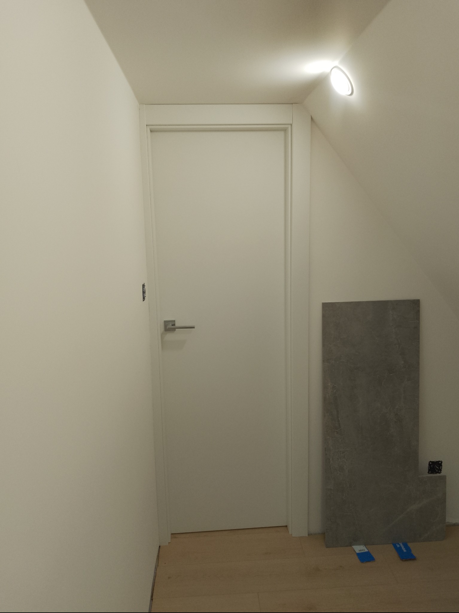 Białe, minimalistyczne drzwi wewnętrzne z klamką, ustawione w jasnym pomieszczeniu ze skośnym sufitem. Obok drzwi leżą płytki podłogowe.