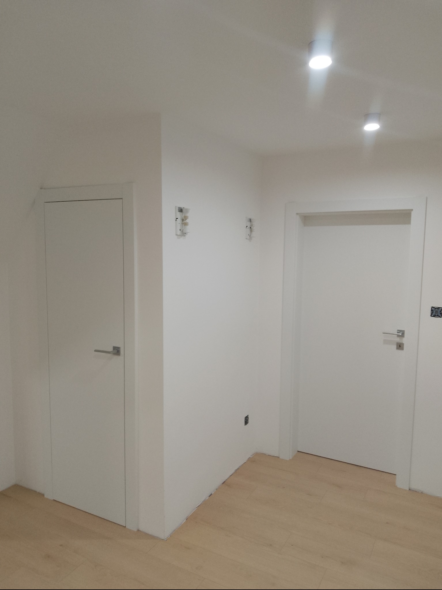 Minimalistyczne wnętrze: dwie białe, pełne drzwi z srebrnymi klamkami, jasna podłoga, białe ściany z włącznikami i haczykami, oświetlenie sufitowe.