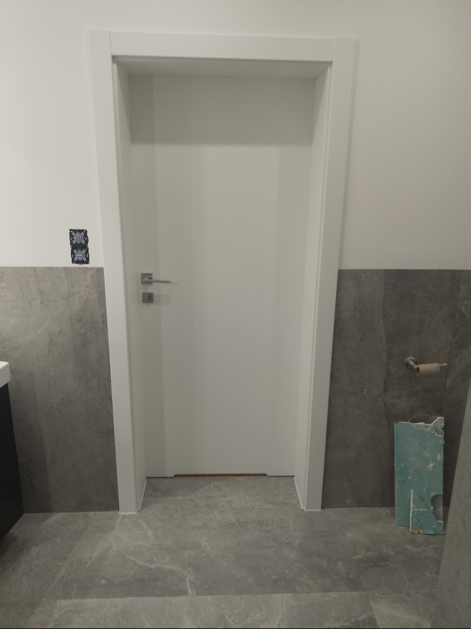 Białe drzwi wewnętrzne z ościeżnicą w łazience z szarymi płytkami. Widoczne gniazdka elektryczne i uchwyt na papier toaletowy. Podłoga wyłożona szarymi płytkami.