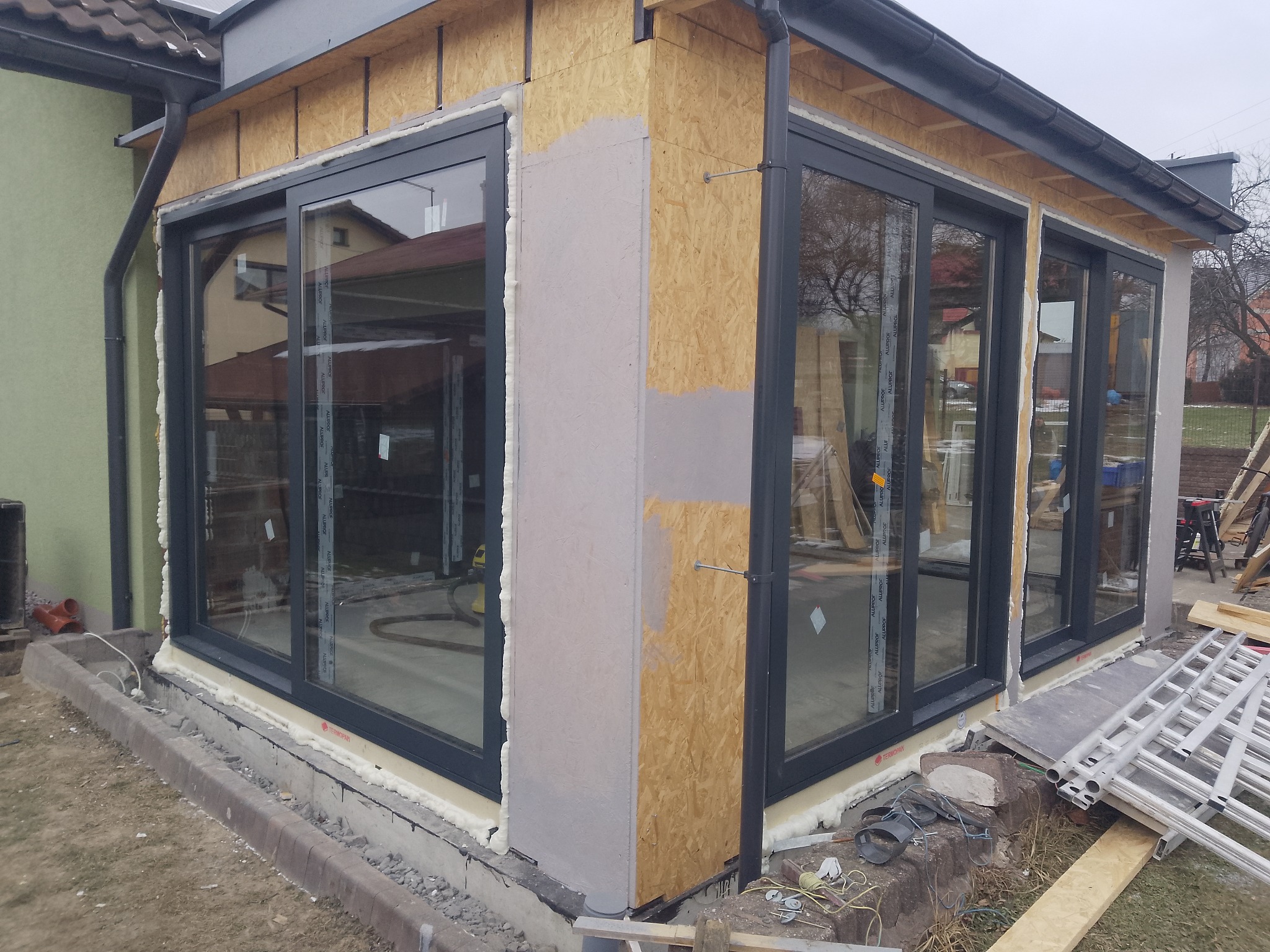 Montaż okien przesuwnych: widok z zewnątrz na szare ramy okienne w trakcie instalacji, z widoczną pianką montażową i elementami konstrukcyjnymi budynku.