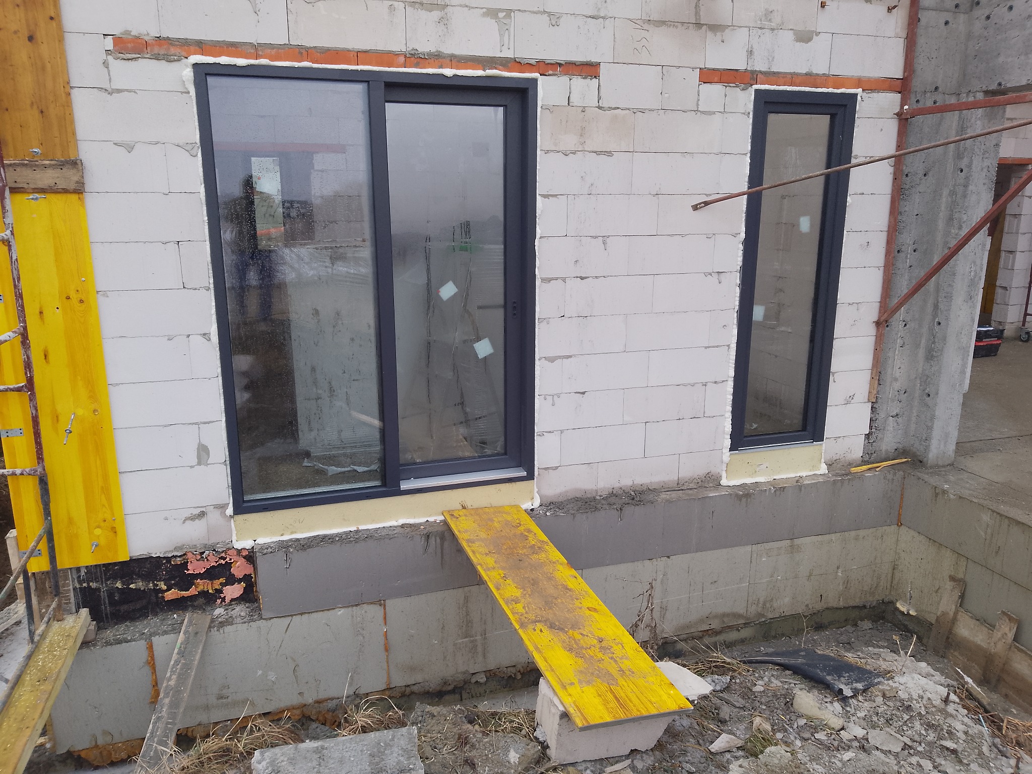 Montaż okien przesuwnych i standardowych w surowym budynku z białego bloczka. Widoczne żółte deski szalunkowe i fragmenty ocieplenia fundamentów.