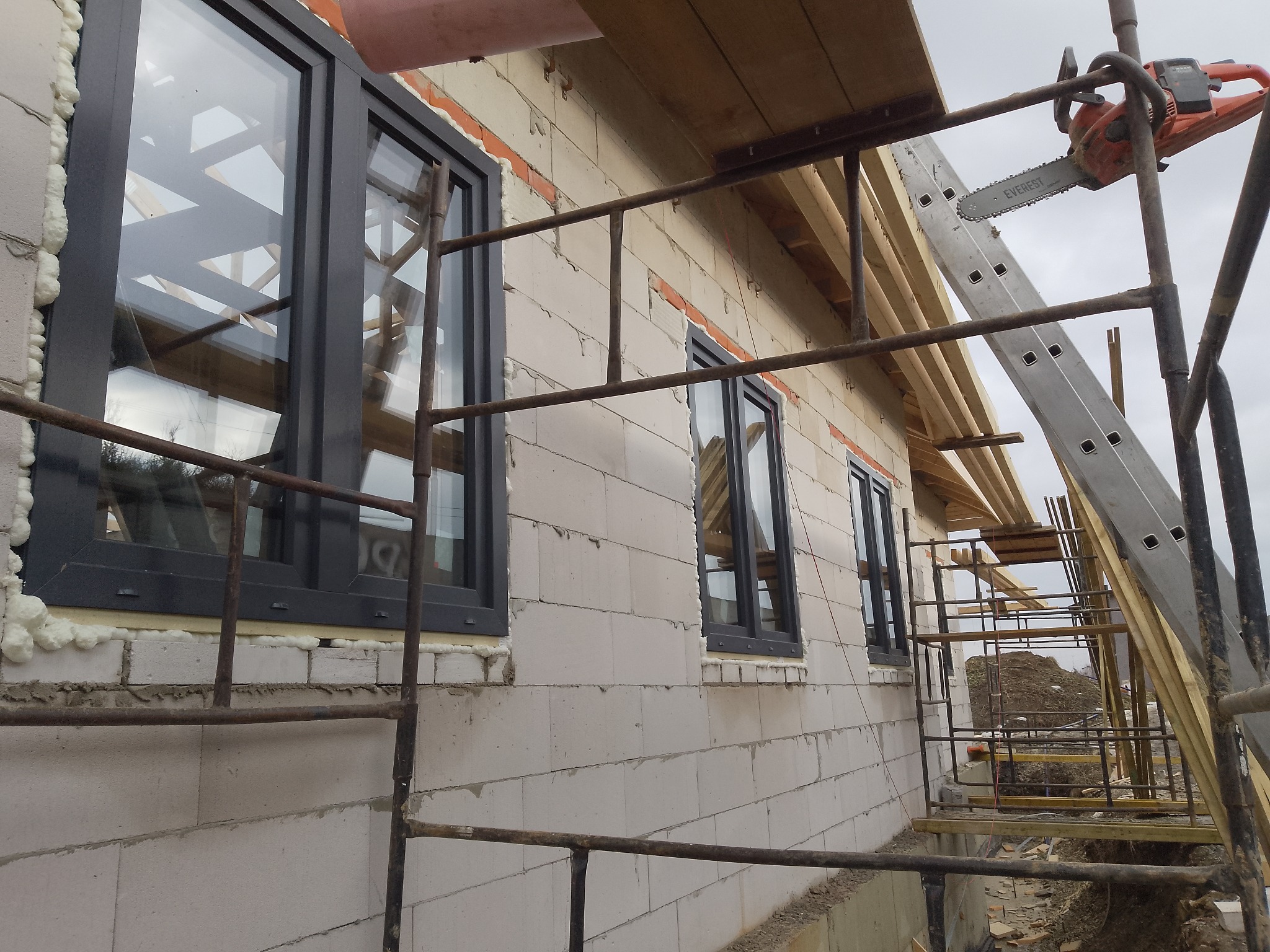 Montaż okien PCV w surowym budynku z białego bloczka. Widoczne rusztowanie, drabina i piła łańcuchowa. Okna w ciemnych ramach, uszczelnione pianką montażową.