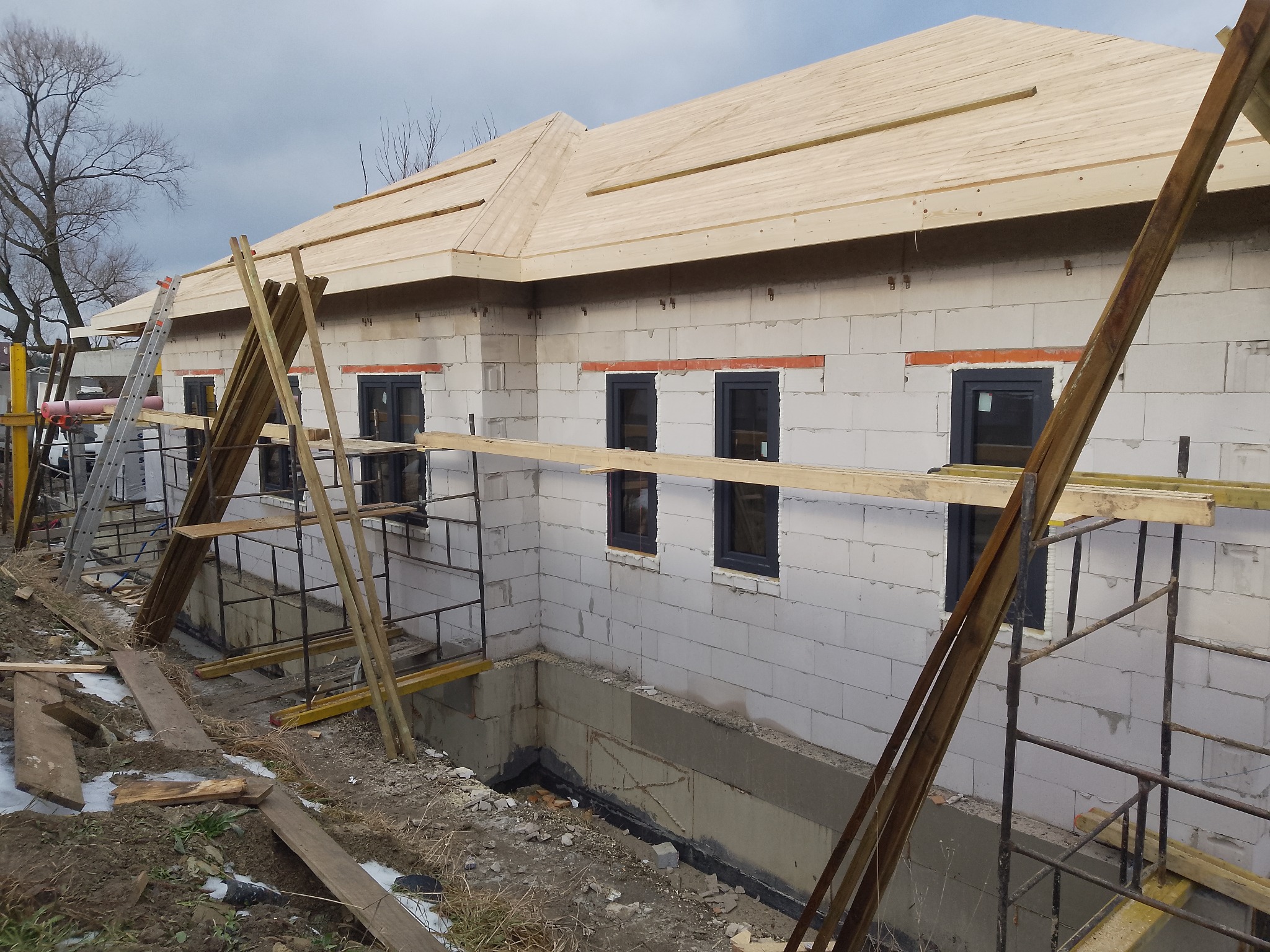 Budowa domu z bloczków: wstawione okna z czarnymi ramami, drewniana konstrukcja dachu w trakcie montażu, rusztowania i materiały budowlane wokół.