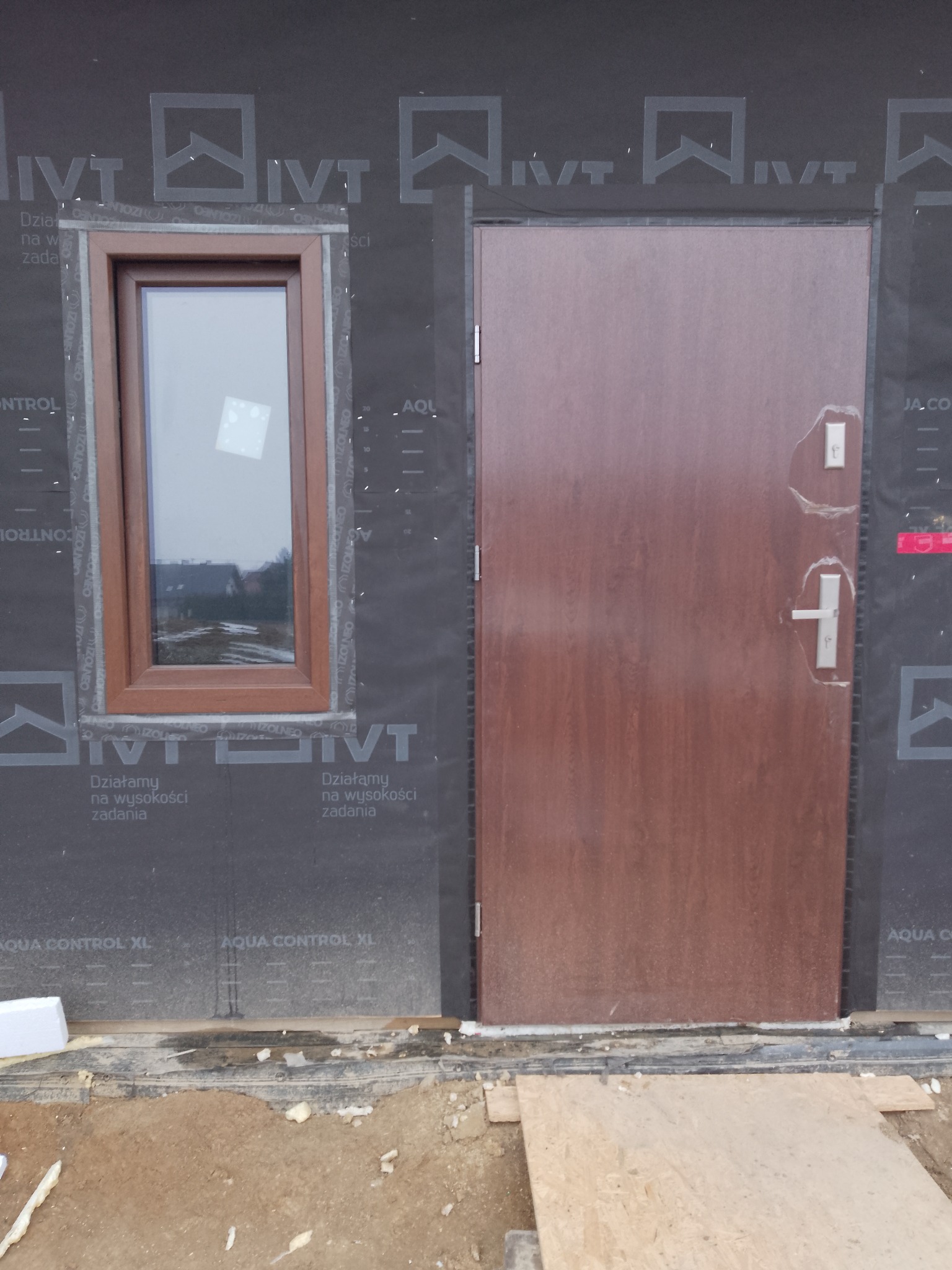 Brązowe drzwi zewnętrzne i okno z brązową ramą na tle czarnej membrany. Widoczne ślady montażu i fragment terenu budowy. Ujęcie z bliska.