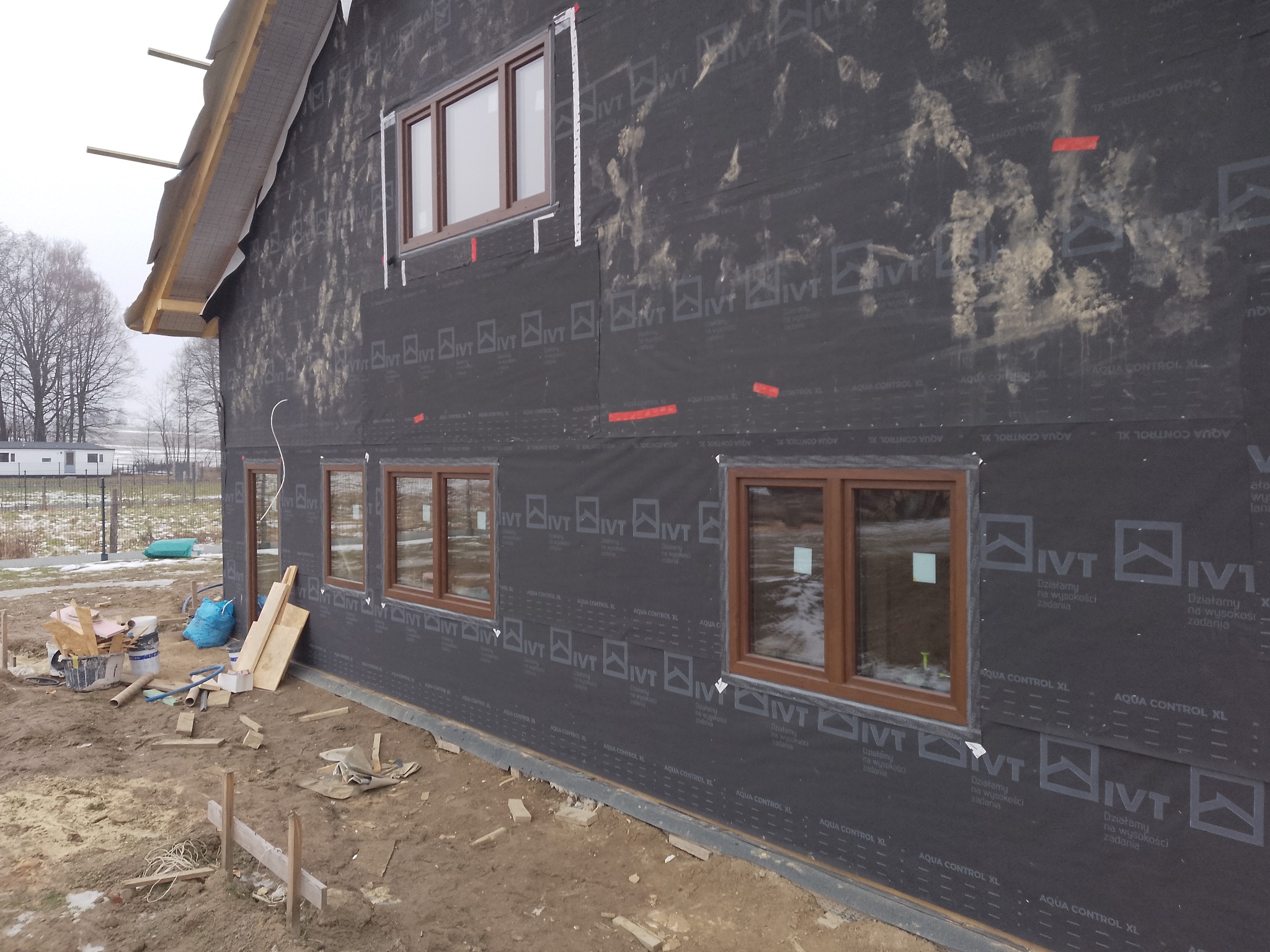 Montaż okien z brązowymi ramami w domu w trakcie budowy, ściany pokryte czarną membraną. Widoczne materiały budowlane i elementy konstrukcyjne.