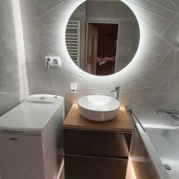 Firma Usługowa Kris Krzysztof Krzysztoń - Nowoczesna łazienka z okrągłym lustrem LED, umywalką nablatową na szafce z szufladami i pralką. Płytki ścienne w geometryczny wzór, podświetlenie pod wanną.