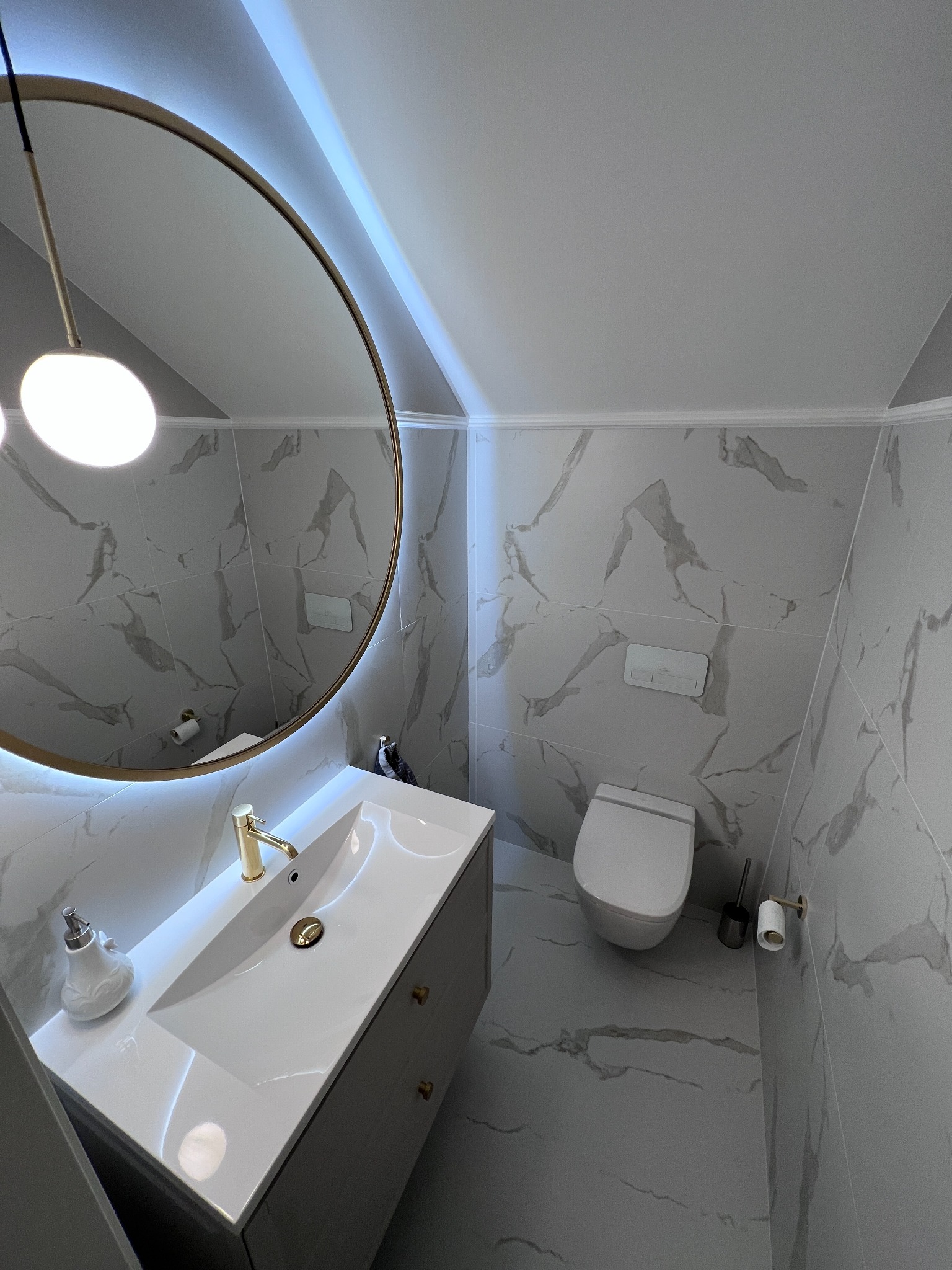 Wykończona łazienka na poddaszu z marmurowymi płytkami, okrągłym lustrem z podświetleniem LED i białą armaturą. Złota bateria umywalkowa i oświetlenie.