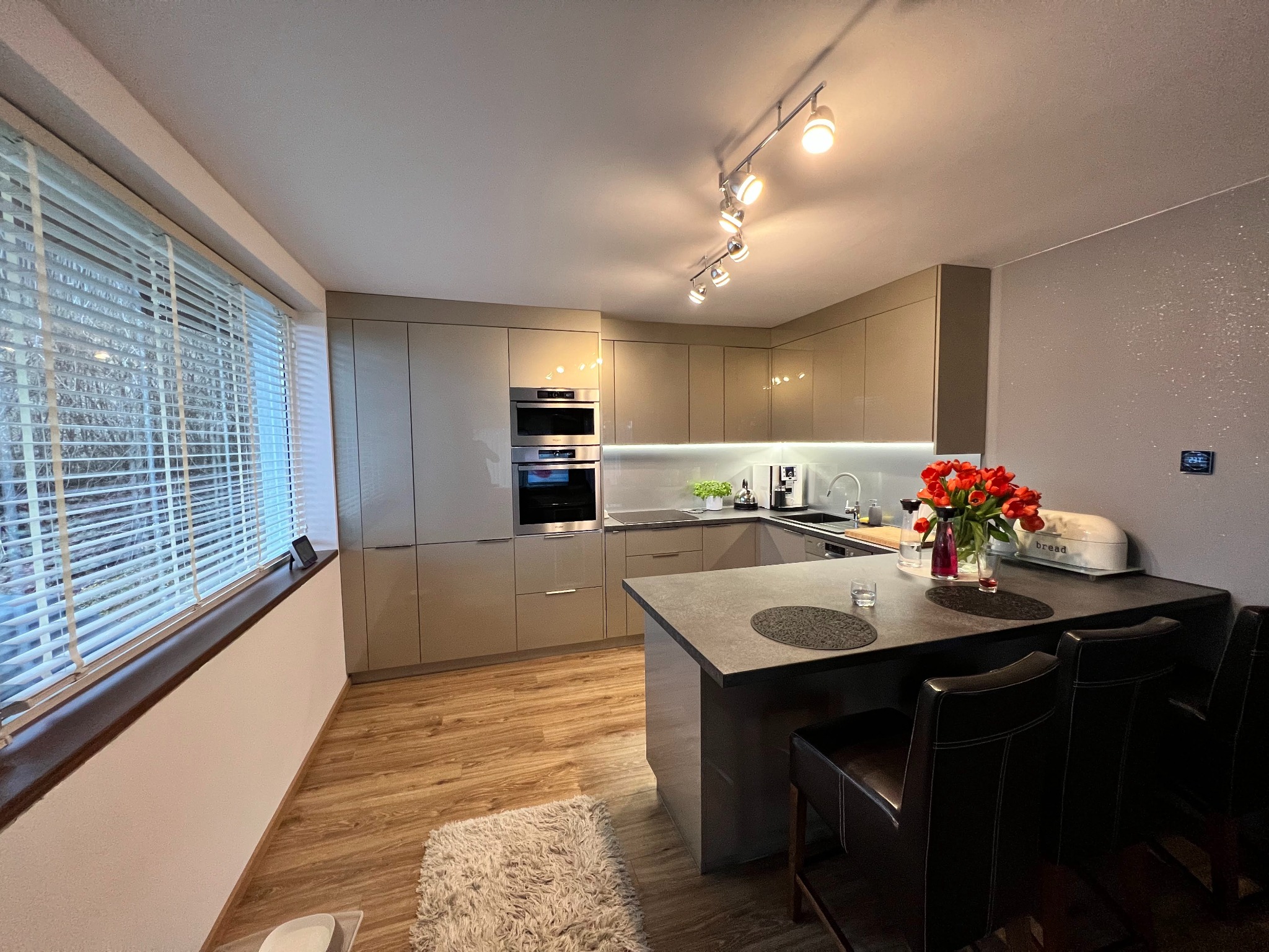 Nowoczesna kuchnia z beżowymi szafkami, wyspą z ciemnym blatem i hokerami. Na blacie kwiaty i chlebak. Parkiet na podłodze, żaluzje w oknie. Oświetlenie punktowe.