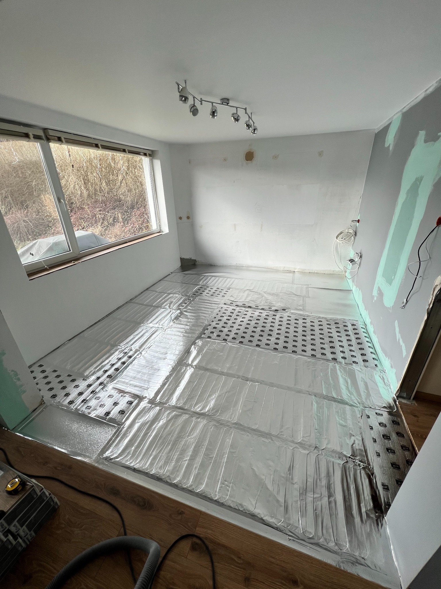 Instalacja ogrzewania podłogowego w pokoju z oknem. Widoczne folie grzewcze na podłodze, przygotowane do dalszych prac wykończeniowych. Ściany częściowo przygotowane do malowania.