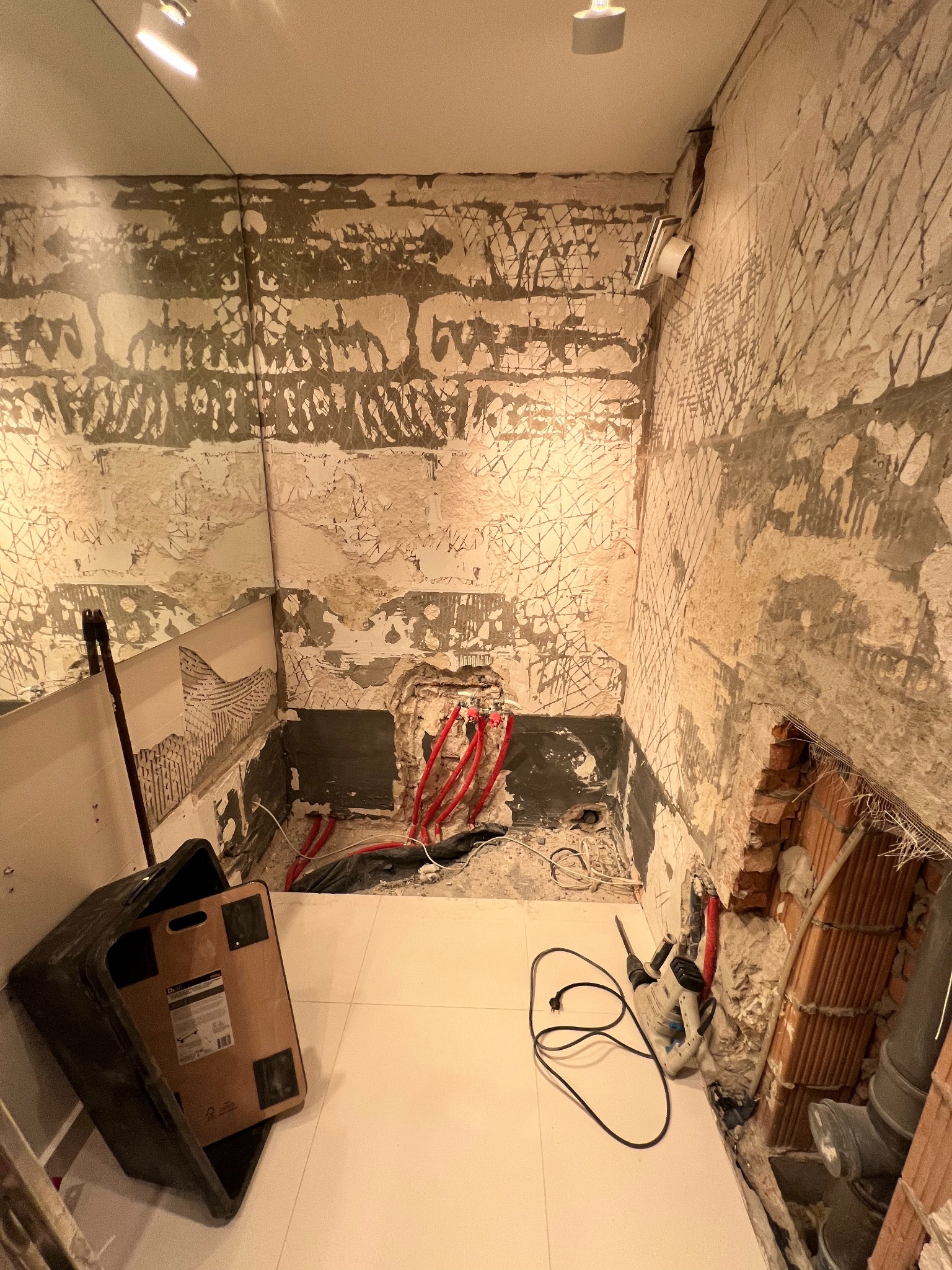 Łazienka w trakcie demontażu płytek, z widocznymi instalacjami i narzędziami. Ściany częściowo skute, ukazujące surowy mur. Nowe, jasne płytki podłogowe.