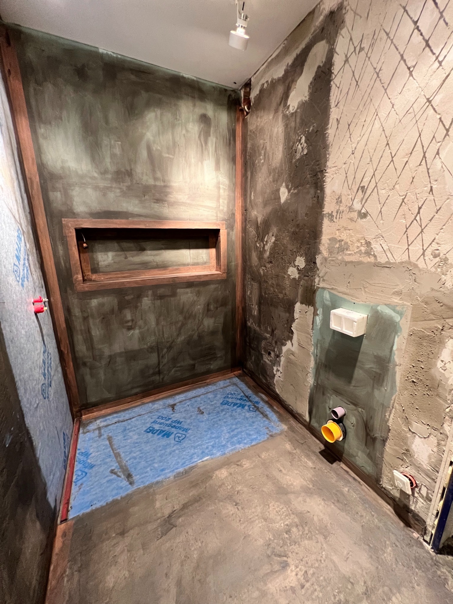 Przygotowanie do wykończenia kabiny prysznicowej: hydroizolacja, obudowa wnęki, instalacja odpływu. Surowe ściany z widocznymi warstwami zaprawy i siatki.