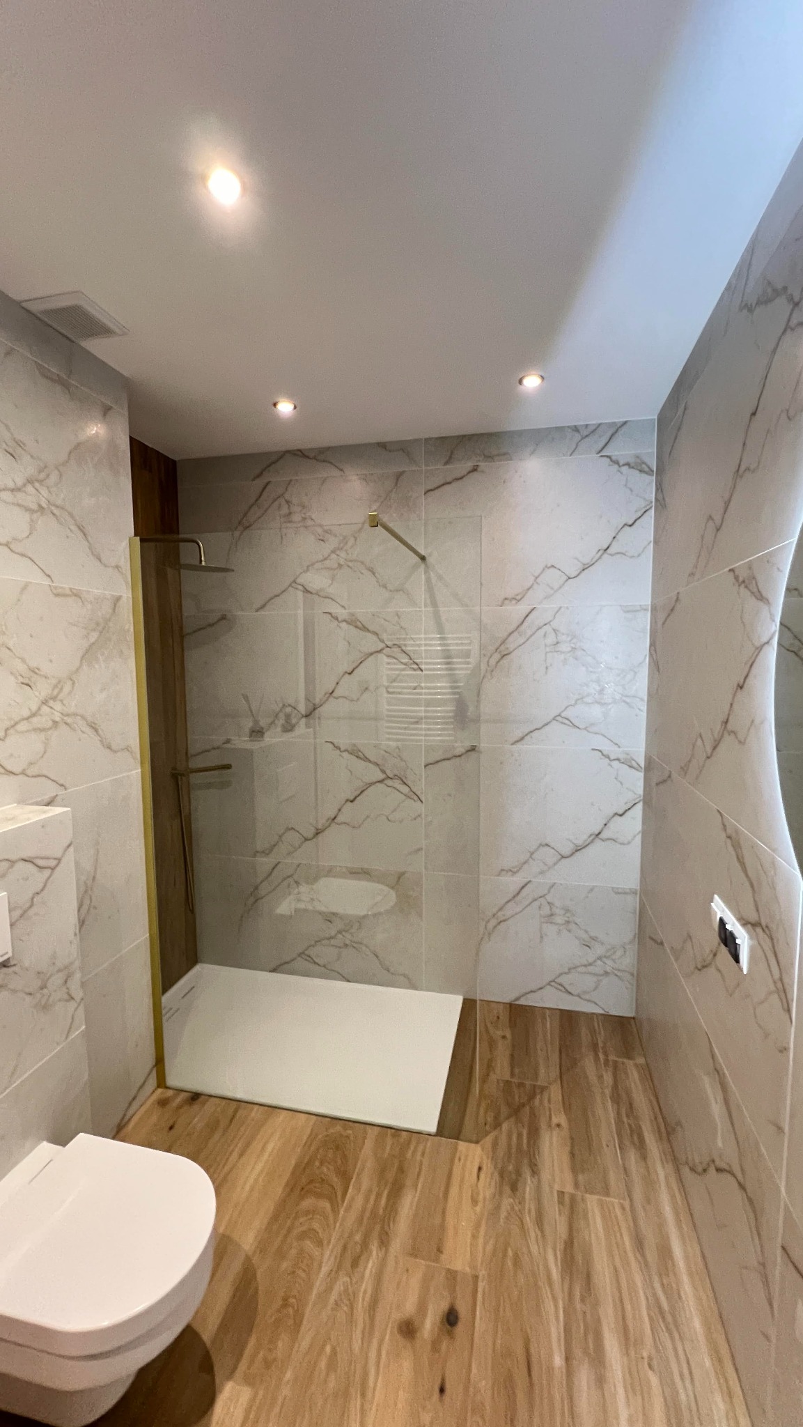 Nowoczesna łazienka z kabiną prysznicową typu walk-in, wykończona płytkami imitującymi marmur i drewnopodobną podłogą. Widoczna toaleta i złote detale.