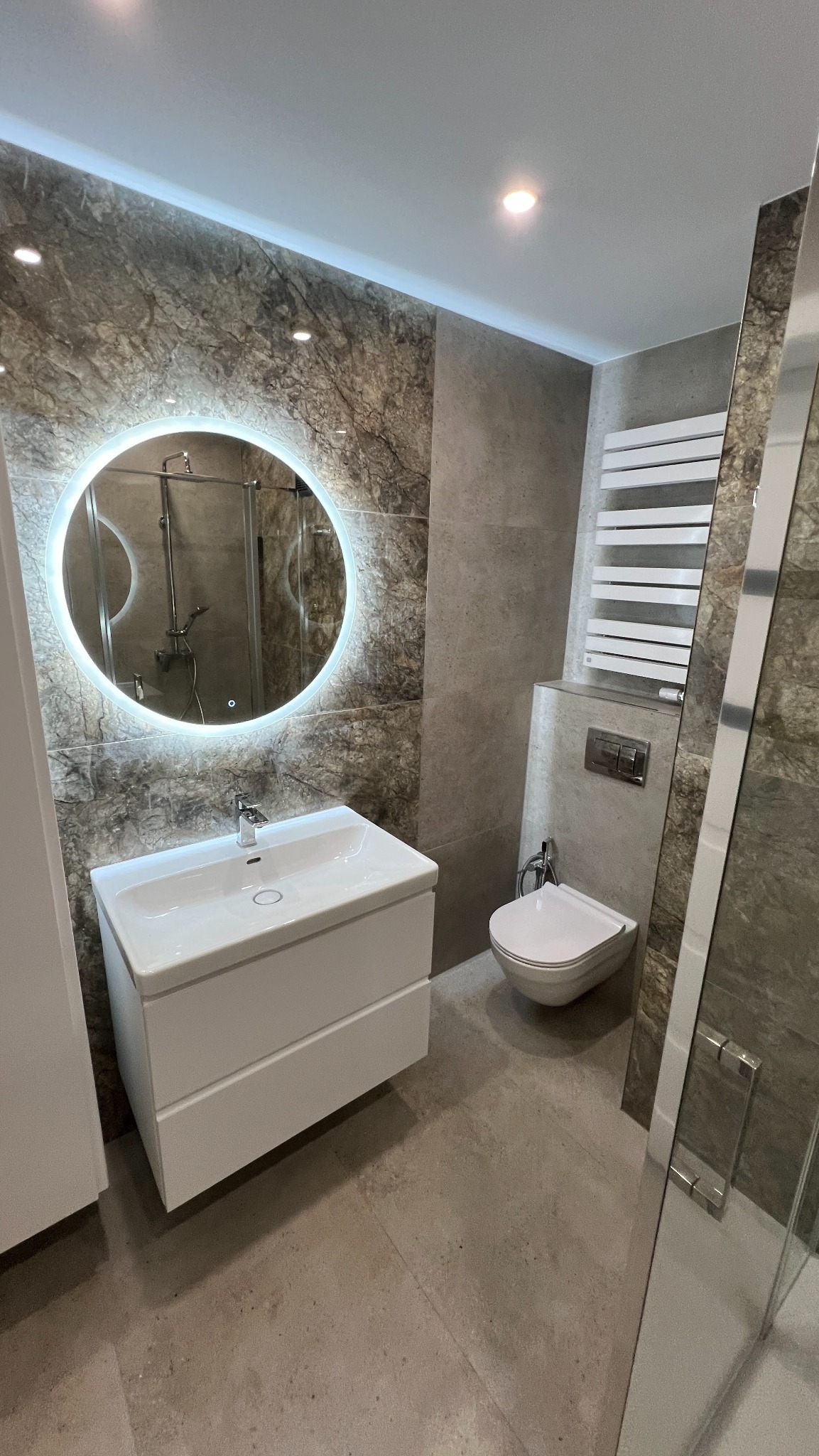 Nowoczesna łazienka z okrągłym lustrem LED, umywalką na szafce i podwieszaną toaletą. Płytki imitujące kamień na ścianach i podłodze. Grzejnik drabinkowy.