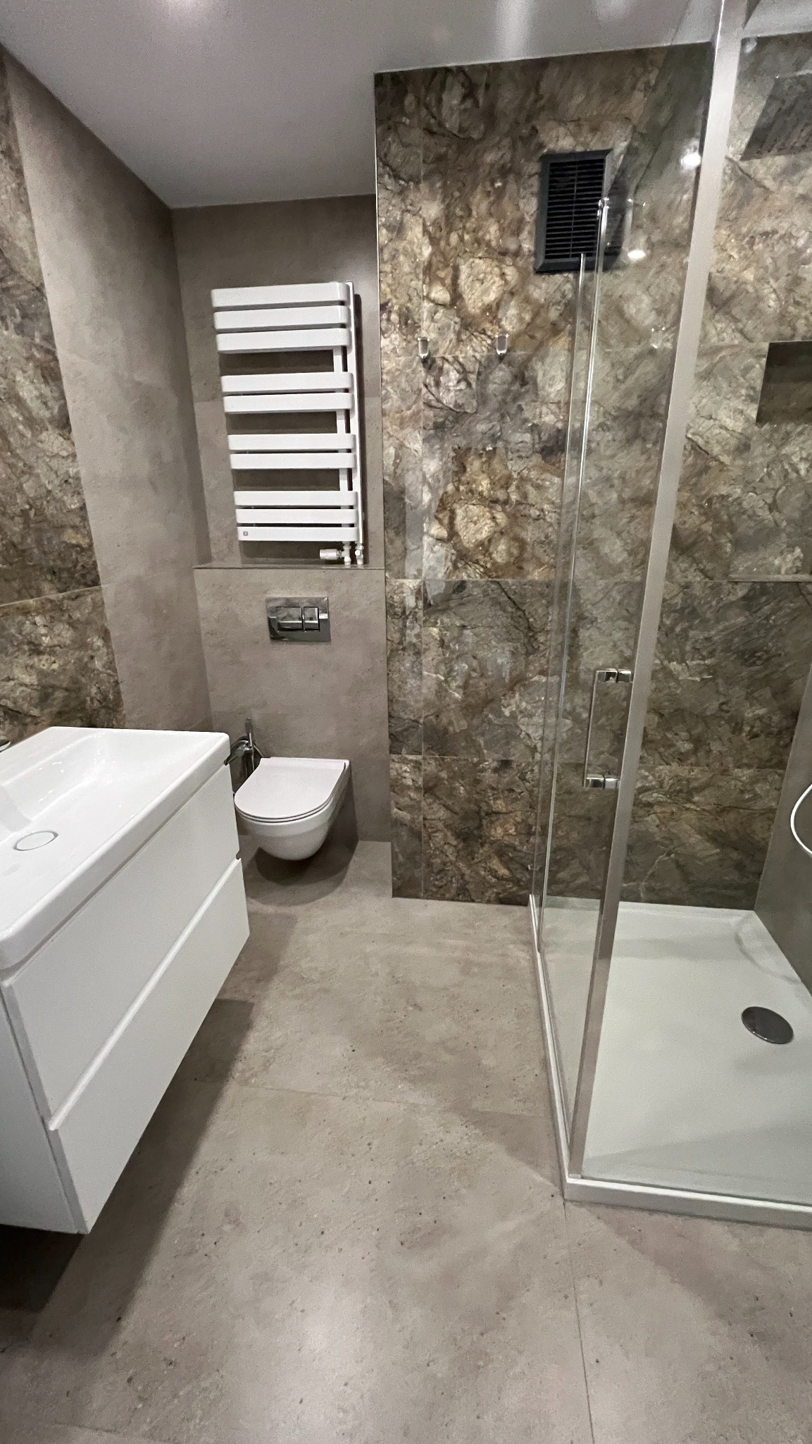 Nowoczesna łazienka z kabiną prysznicową, umywalką z szafką i grzejnikiem drabinkowym. Ściany wykończone płytkami imitującymi marmur. Minimalistyczny design.