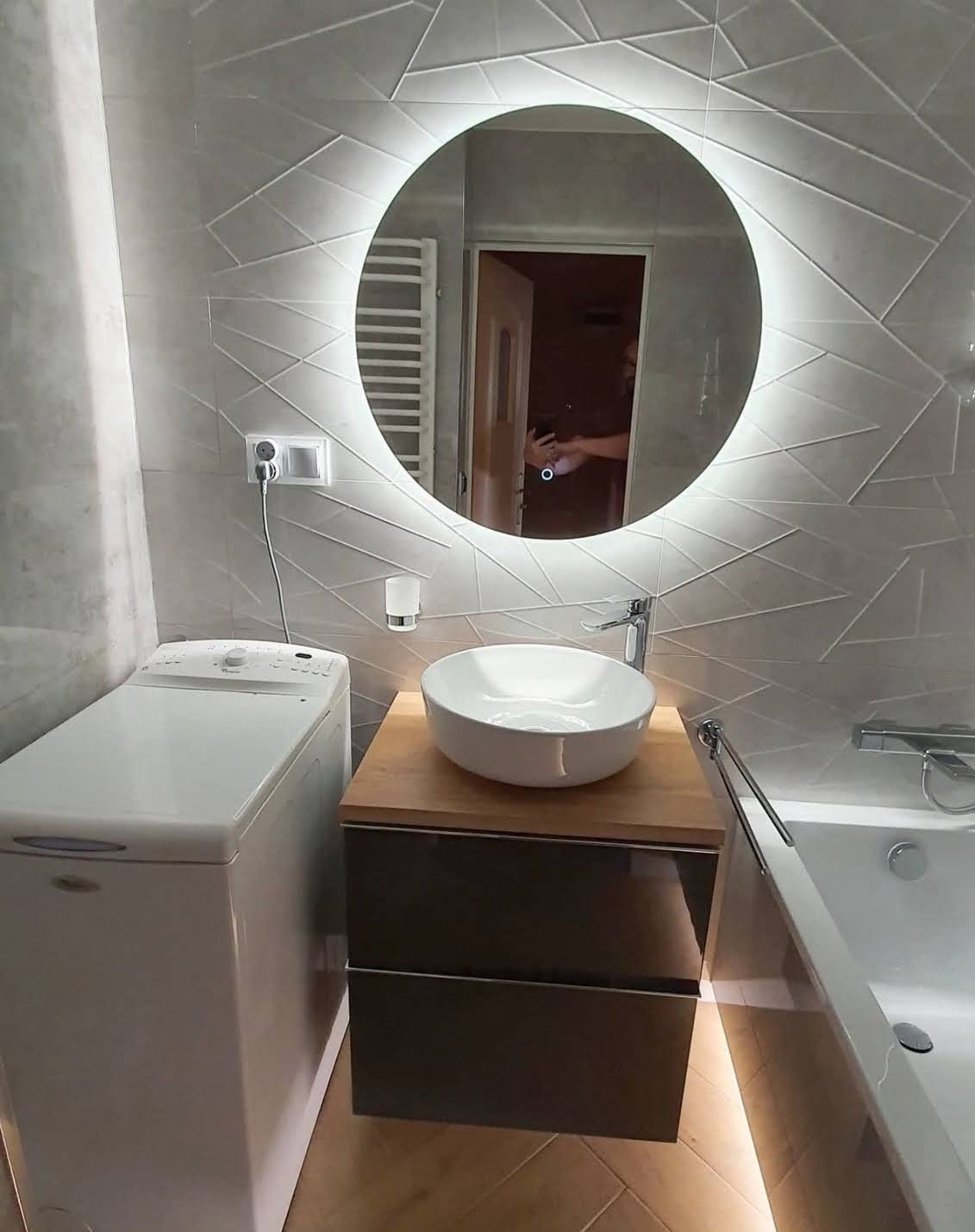 Nowoczesna łazienka z okrągłym lustrem LED, umywalką nablatową na szafce z szufladami i pralką. Płytki ścienne w geometryczny wzór, podświetlenie pod wanną.