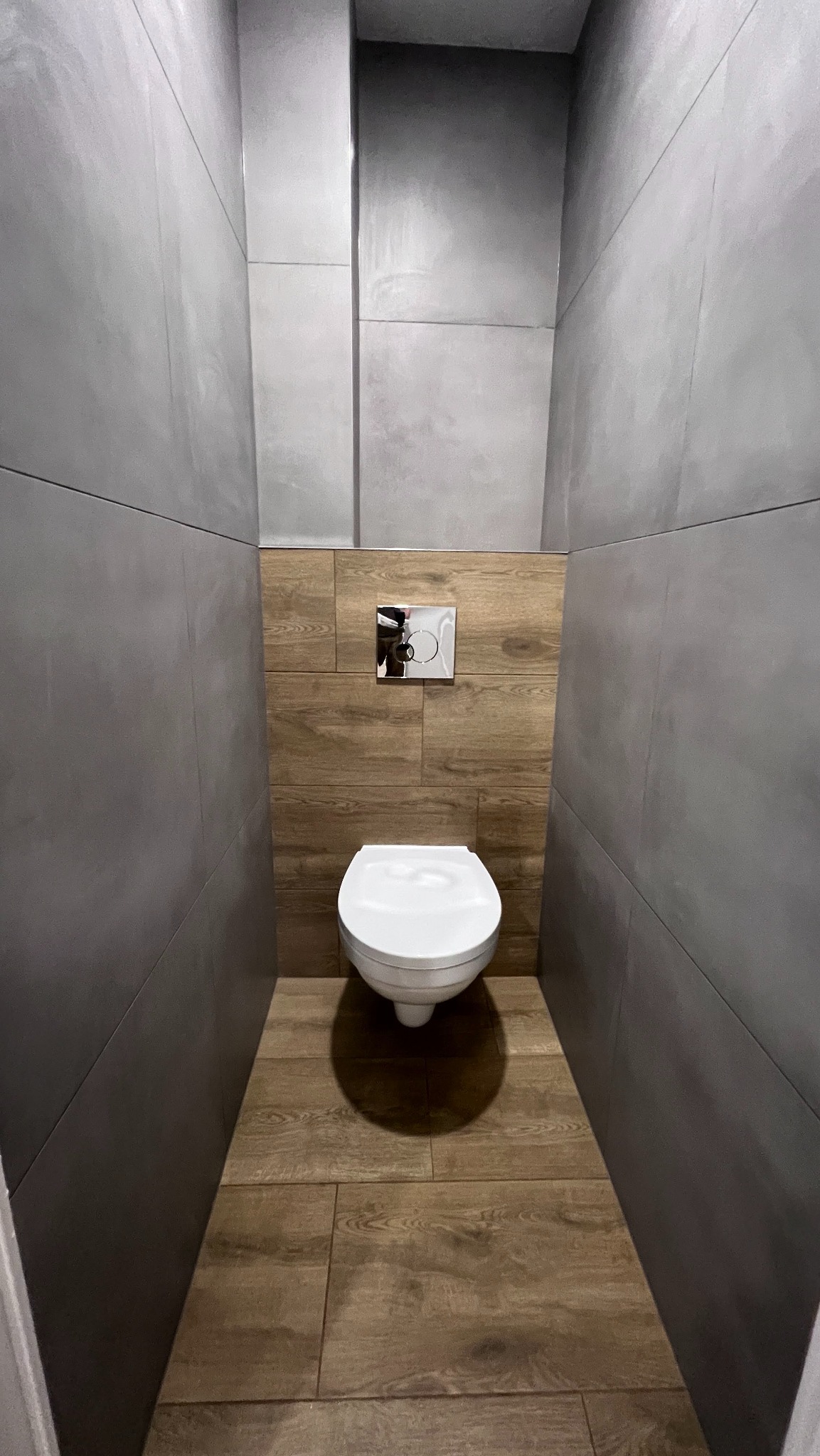 Wąska toaleta z szarymi płytkami ściennymi i drewnopodobną podłogą. Podwieszana muszla klozetowa z chromowanym przyciskiem spłukującym. Minimalistyczny design.