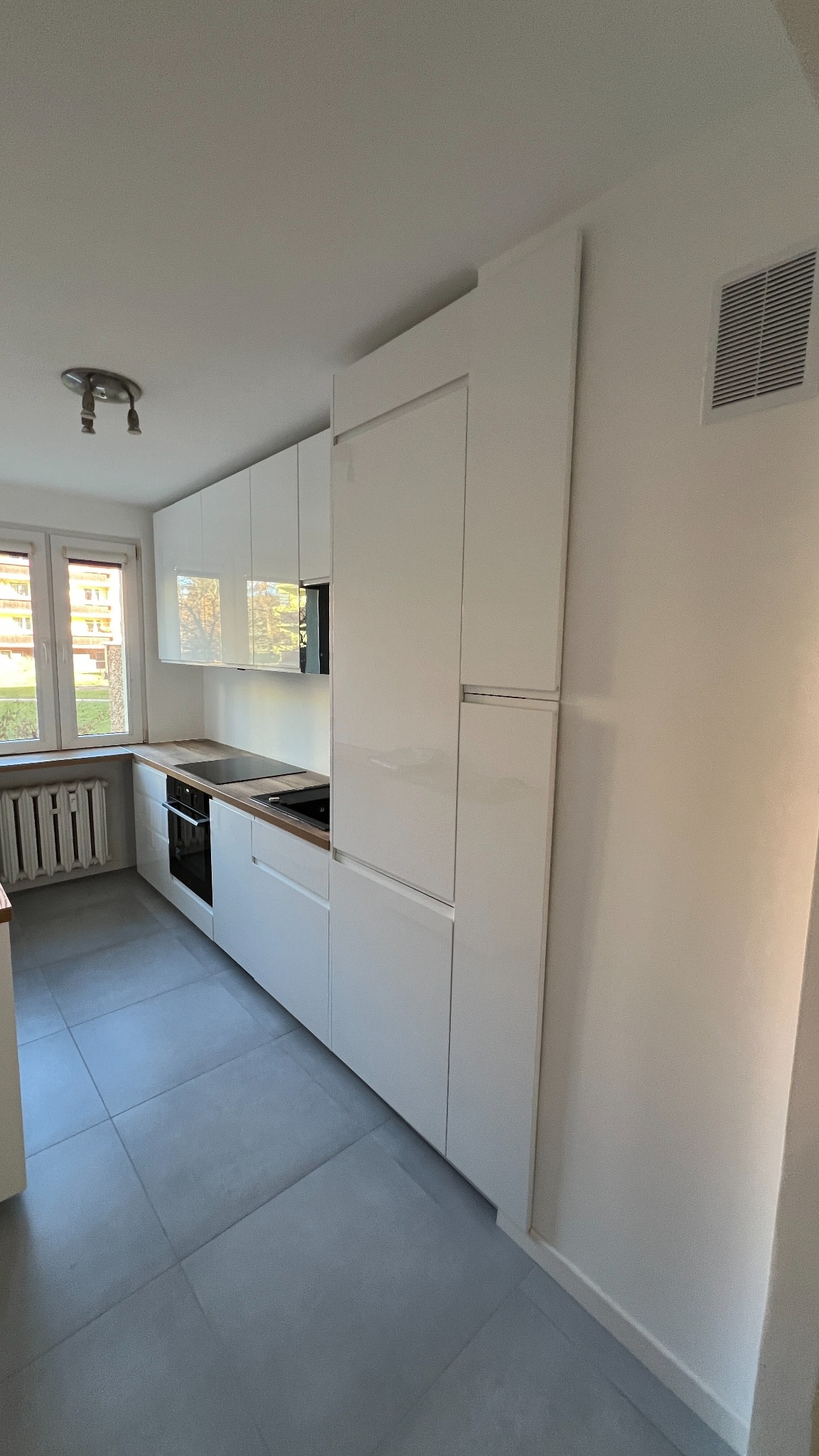 Biała, nowoczesna kuchnia z szarymi płytkami podłogowymi i drewnianym blatem. Widok na okno z grzejnikiem. Minimalistyczny design, jasne wnętrze.