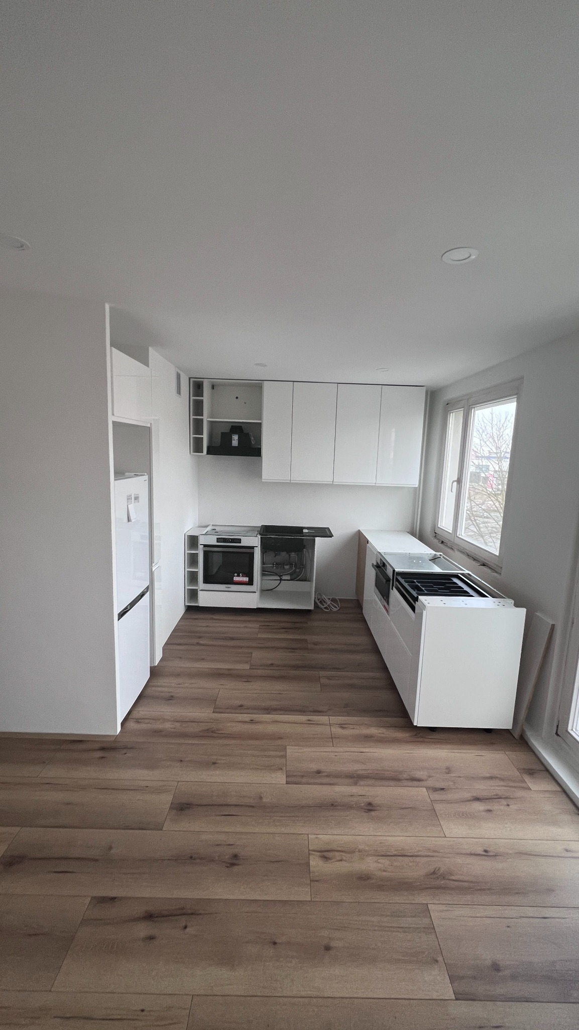 Jasna kuchnia w trakcie montażu: białe szafki wiszące i stojące, drewnopodobna podłoga, okno z widokiem na zewnątrz. Widoczne niedokończone elementy i brakujące blaty.