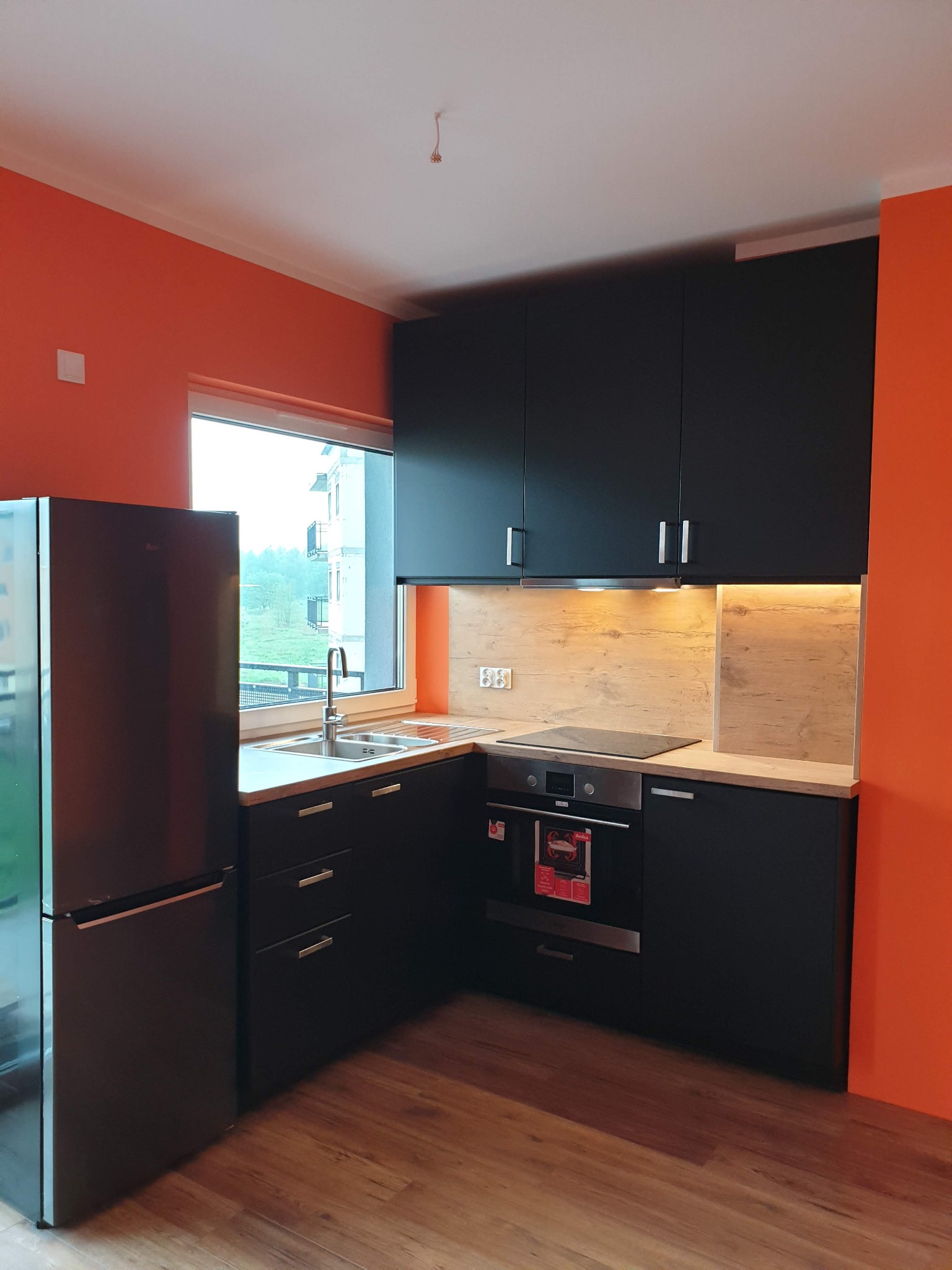 Nowoczesna kuchnia z czarnymi meblami i pomarańczowymi ścianami. Widoczny zlew, płyta indukcyjna i lodówka. Jasne drewno na blacie i podświetlenie LED dodają ciepła.