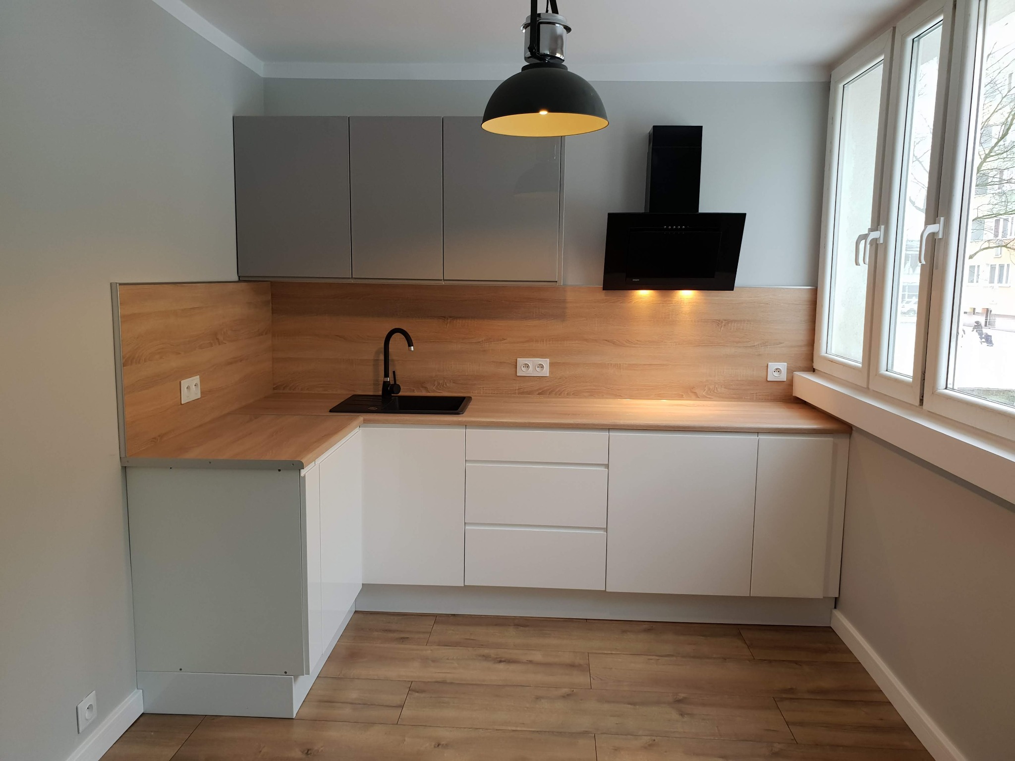 Nowoczesna kuchnia w odcieniach szarości i drewna. Białe fronty szafek dolnych, szare górne. Czarny okap i lampa. Blat drewnopodobny. Okno z widokiem na zewnątrz.