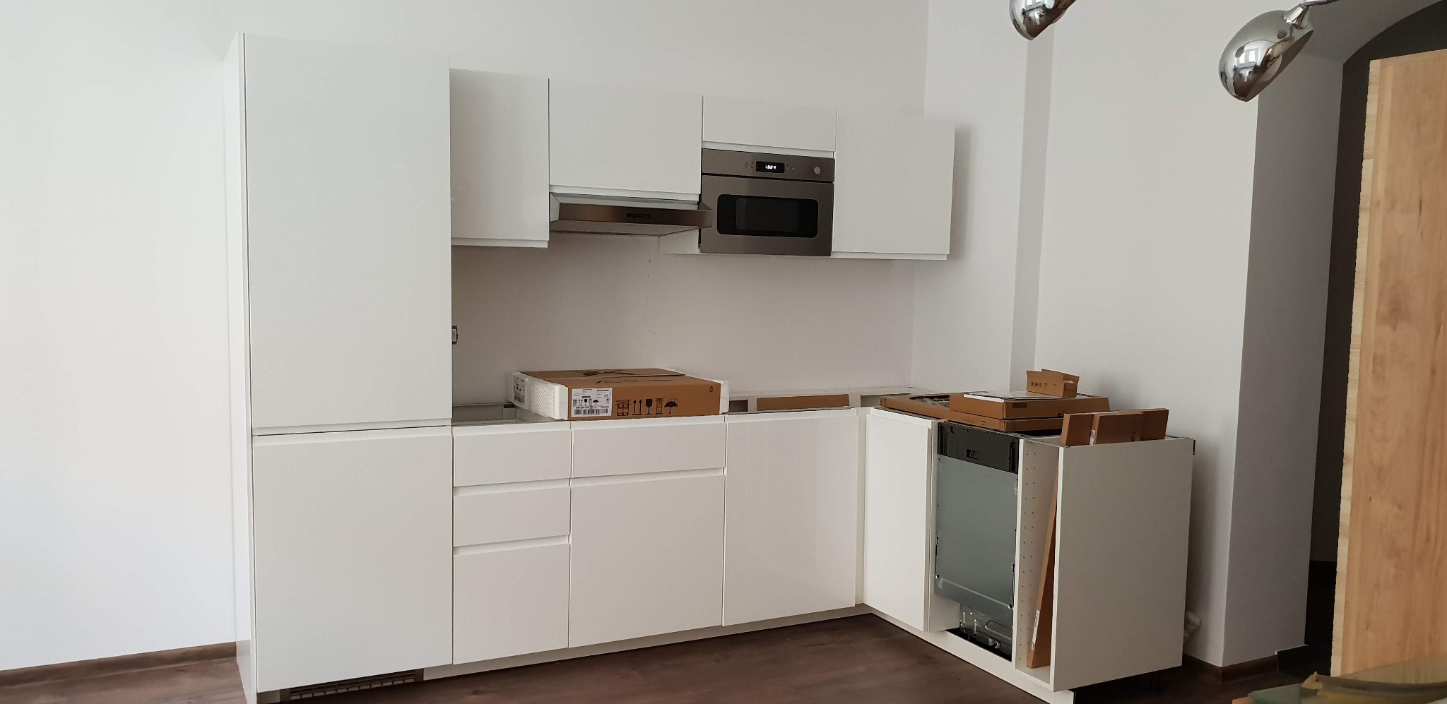 Biała, nowoczesna kuchnia w trakcie montażu. Widoczne szafki, piekarnik, okap i zmywarka w zabudowie. Na blacie kartonowe opakowania. Podłoga z paneli.