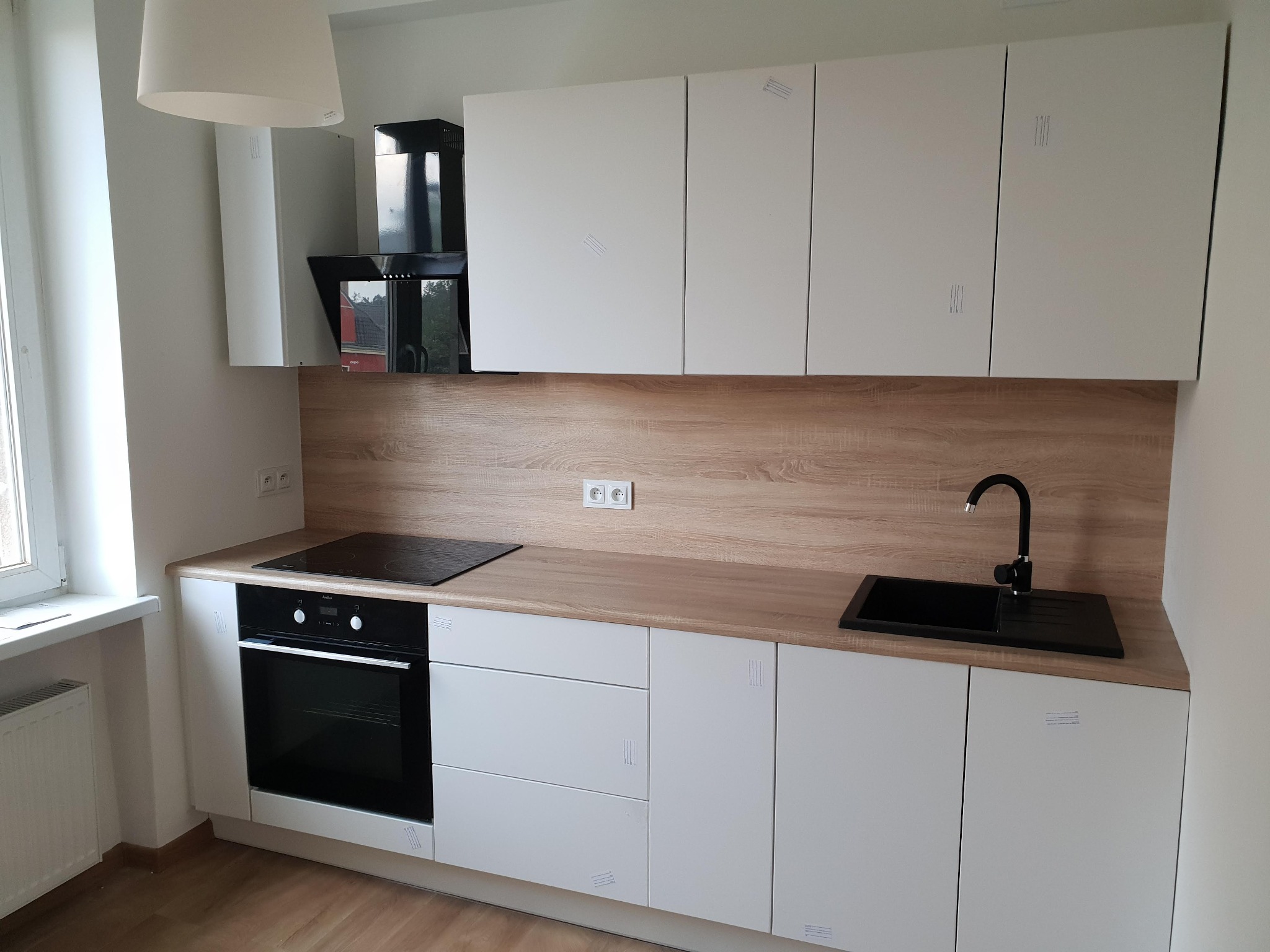 Jasna kuchnia z białymi szafkami i drewnianym blatem. Nowoczesny design z czarnym zlewem i płytą grzewczą. Minimalistyczny styl, świeżo po montażu, widoczne zabezpieczenia.