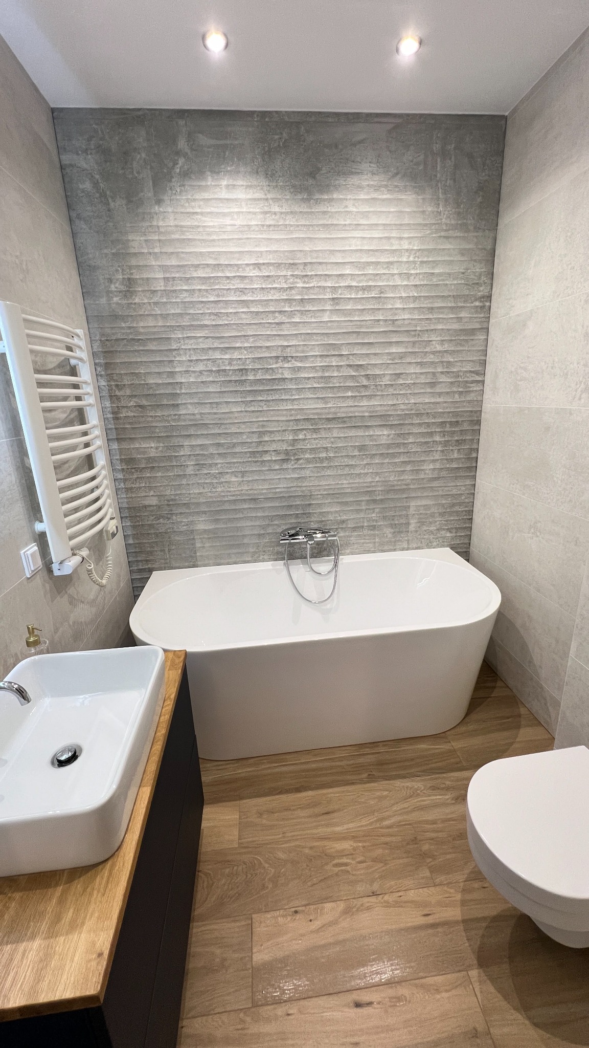 Nowoczesna łazienka z wolnostojącą wanną, umywalką nablatową i grzejnikiem drabinkowym. Szare płytki na ścianach i drewnopodobna podłoga tworzą spójną całość.