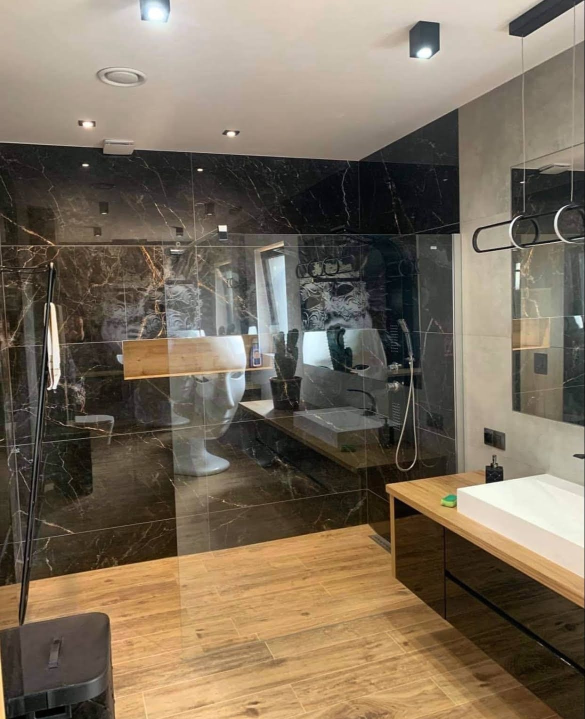 Elegancka łazienka z czarnymi płytkami imitującymi marmur, drewnopodobną podłogą i szklaną kabiną prysznicową. Umywalka nablatowa na szafce z drewnianym blatem.