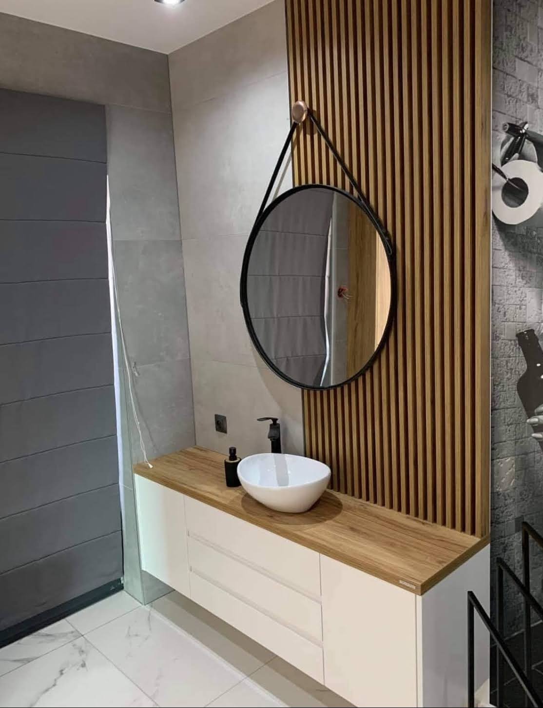 Nowoczesna łazienka z drewnianymi lamelami, okrągłym lustrem na pasku i białą szafką z drewnianym blatem. Minimalistyczny design, szare płytki i marmurowa podłoga.