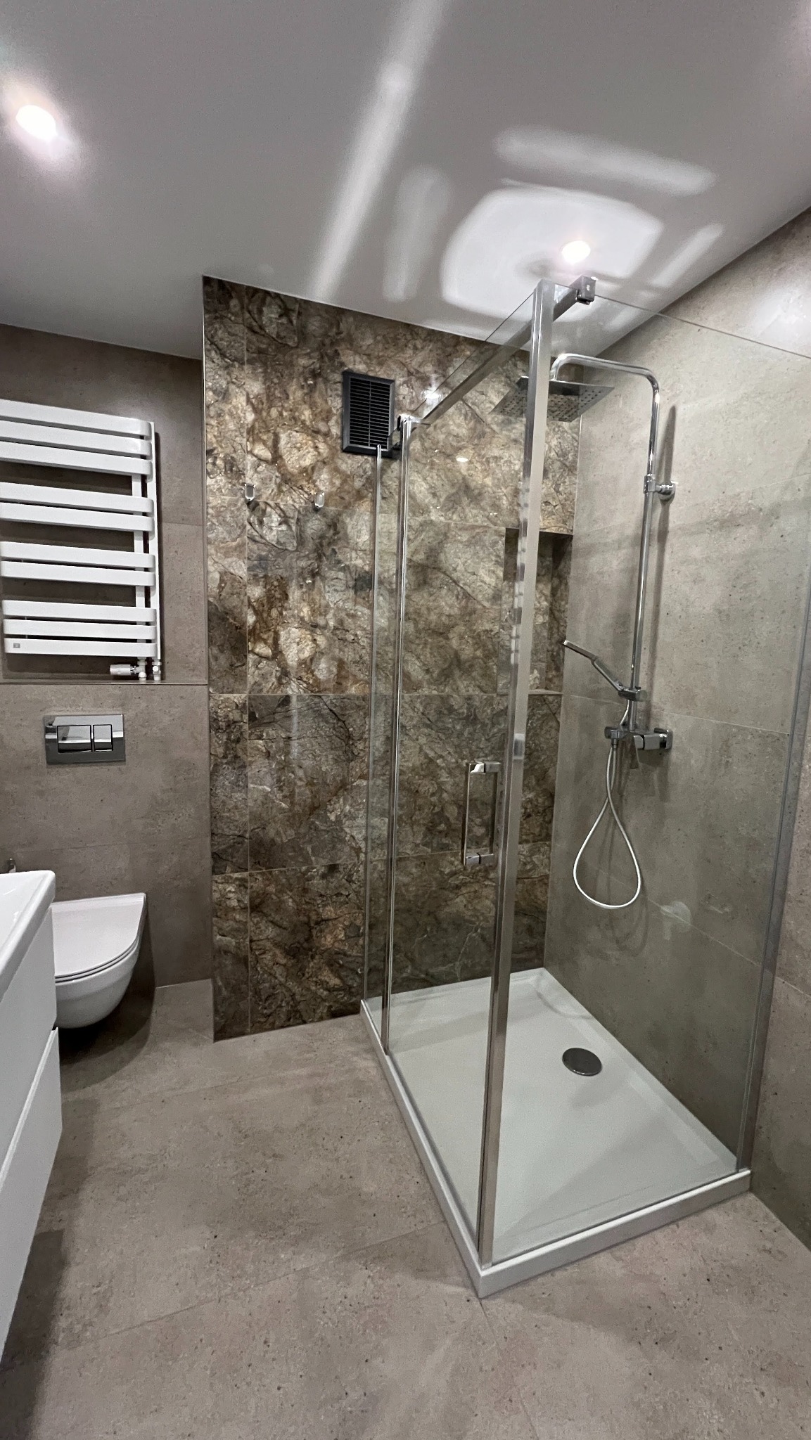 Nowoczesna kabina prysznicowa z marmurową ścianą w wykończonej łazience. Widoczny grzejnik drabinkowy i toaleta. Minimalistyczny design, szare płytki podłogowe.