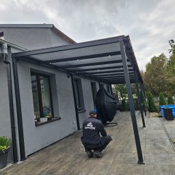 SpecStal Dawid Kędzierski - Nowoczesna, grafitowa pergola z oświetleniem LED, przylegająca do domu. Pracownik firmy instalacyjnej w trakcie pracy. W tle basen i zieleń ogrodu.