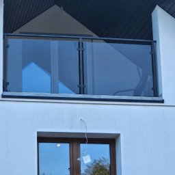 SpecStal Dawid Kędzierski - Nowoczesna balustrada szklana z czarnymi profilami na balkonie domu z białymi ścianami. Widoczny fragment okna i lampa zewnętrzna. Minimalistyczny design.