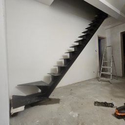 SpecStal Dawid Kędzierski - Nowoczesne, metalowe schody ażurowe w surowym wnętrzu. Widoczne narzędzia i drabina, co sugeruje etap montażu. Minimalistyczny design, czarne elementy na tle białej ściany.