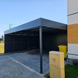 SpecStal Dawid Kędzierski - Nowoczesna, czarna wiata garażowa z metalowymi panelami, przylegająca do budynku z żółtym licznikiem gazu i koszem na śmieci. Kostka brukowa na podjeździe.