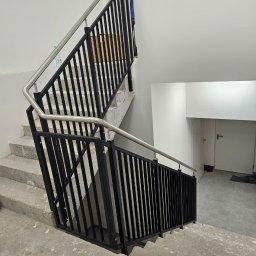 SpecStal Dawid Kędzierski - Nowoczesna balustrada schodowa z czarnymi, pionowymi elementami i stalową poręczą, zamontowana w surowym, betonowym wnętrzu klatki schodowej.