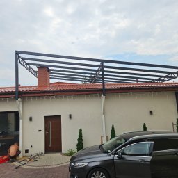 SpecStal Dawid Kędzierski - Montaż metalowej pergoli przy domu jednorodzinnym. Widoczny pracownik układający kostkę brukową. Częściowo widoczny samochód osobowy.