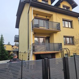 SpecStal Dawid Kędzierski - Żółty dom z balkonami zabezpieczonymi poziomymi, metalowymi panelami. W tle drabina oparta o balkon. Na pierwszym planie fragment ogrodzenia z paneli.