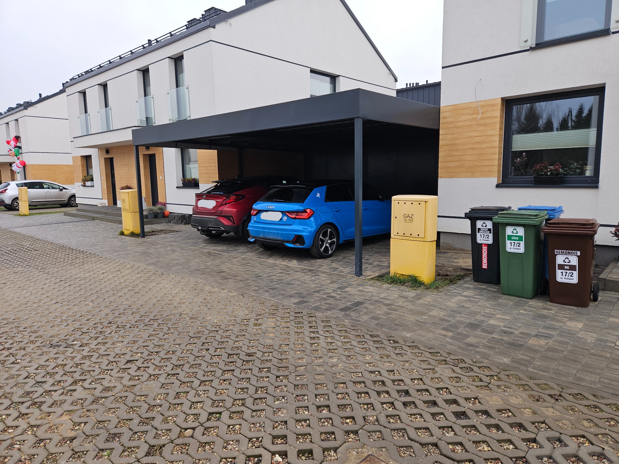 Nowoczesna, grafitowa wiata garażowa z dwoma samochodami (czerwonym i niebieskim) na posesji z kostki ażurowej. W tle budynki mieszkalne.