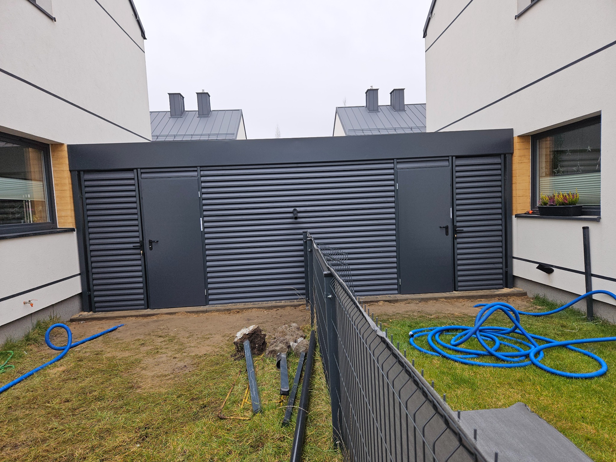 Nowoczesna, antracytowa zabudowa garażowa z lamelami, wkomponowana między budynki. Widoczny fragment ogrodzenia panelowego i węże ogrodowe na trawniku.